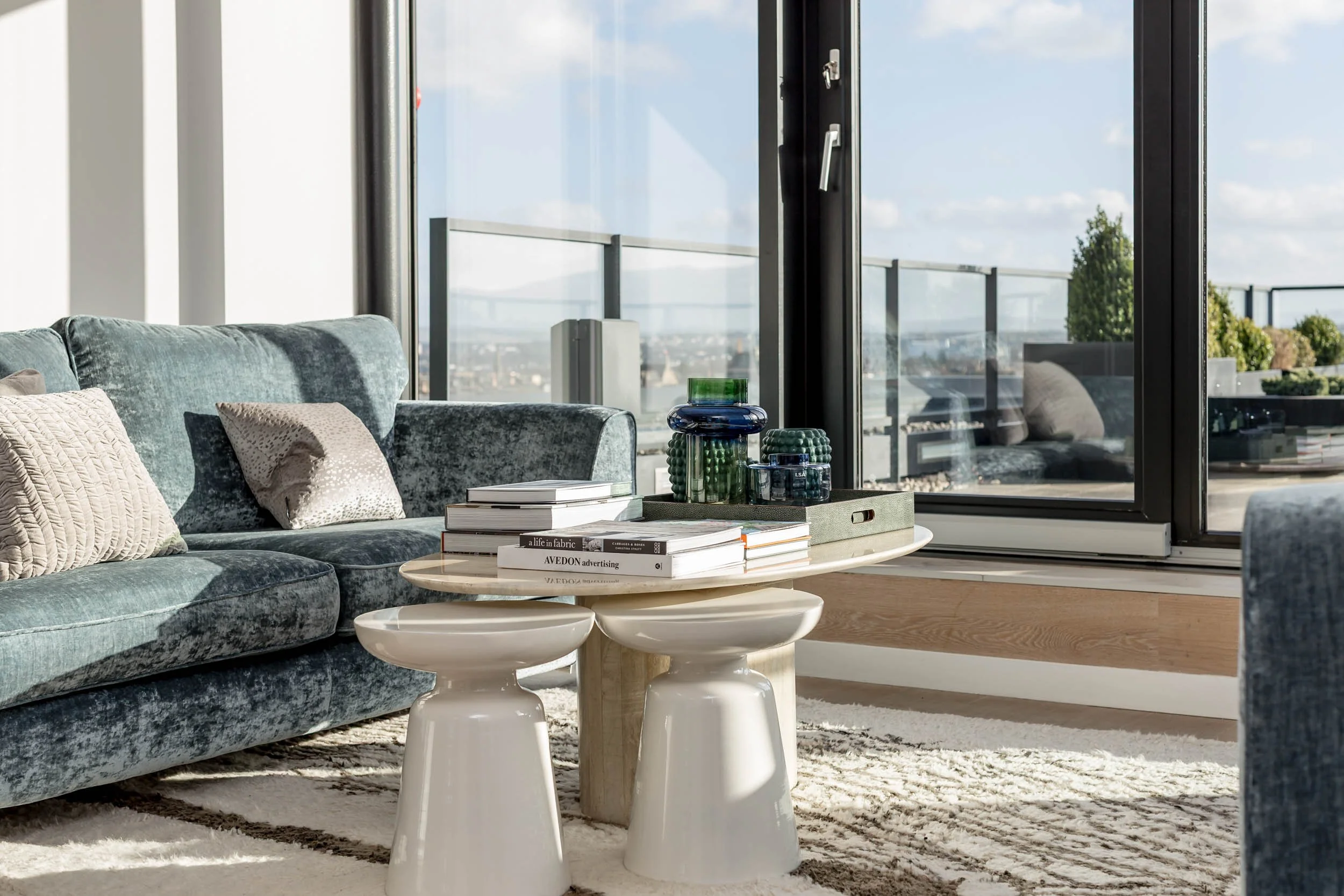 Modern living room with a blue velvet sofa, beige pillows, a white marble coffee table, some decorative glass items, the room has large glass doors opening to a balcony with city views, and a rug on the floor.