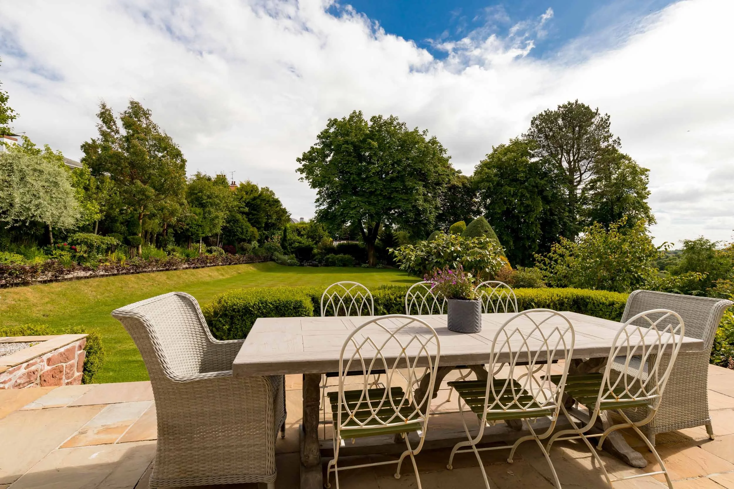 Outdoor patio with a white wooden dining table surrounded by six white metal chairs and two wicker armchairs, with a potted plant on the table, overlooking a lush garden with trees, shrubs, and a well-maintained lawn under a partly cloudy sky.