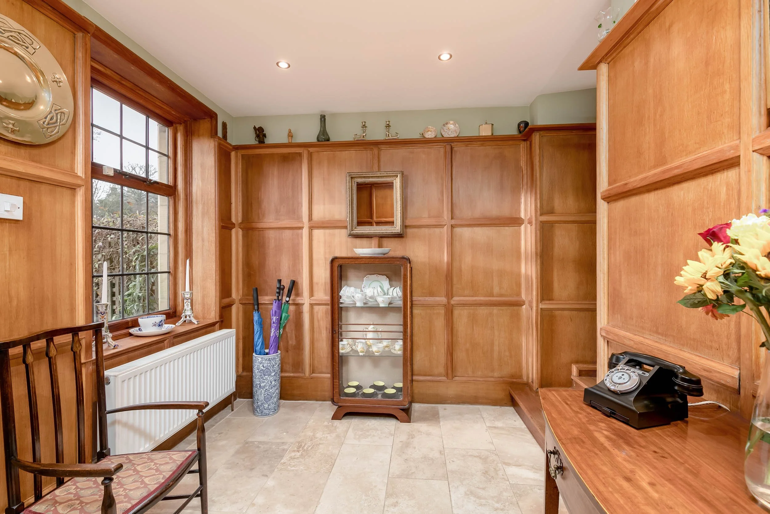 A room with wooden wall paneling, a window with candles and a teacup on the sill, a small display cabinet with china, an armchair, umbrella stand, a rotary phone on a wooden desk, and a vase of flowers.