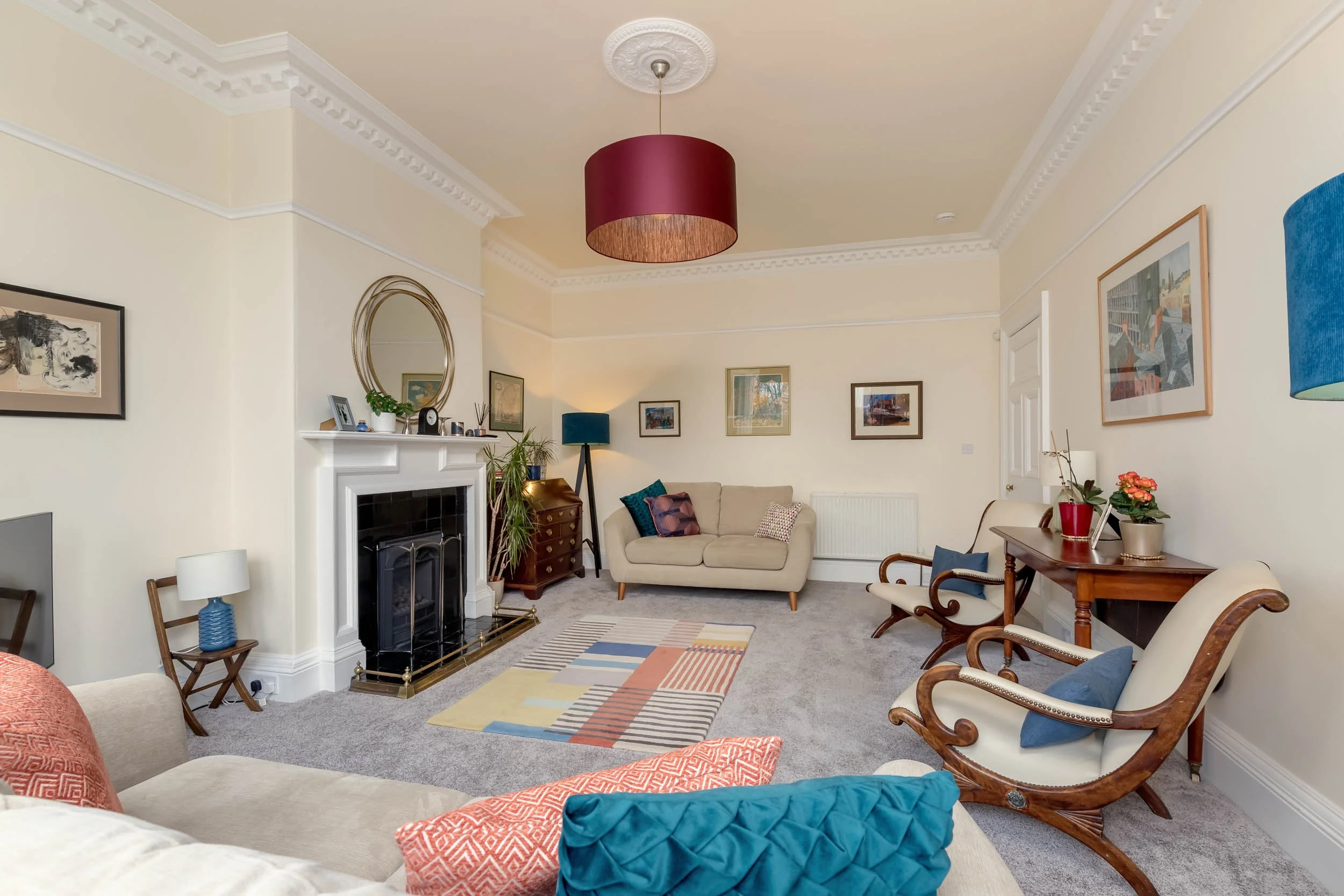 Bright living room with cream-colored walls, ornate ceiling molding, and a gray carpet. Features a white fireplace with a round mirror and decorative items on the mantle, a variety of framed artwork, traditional and modern furniture including a cream