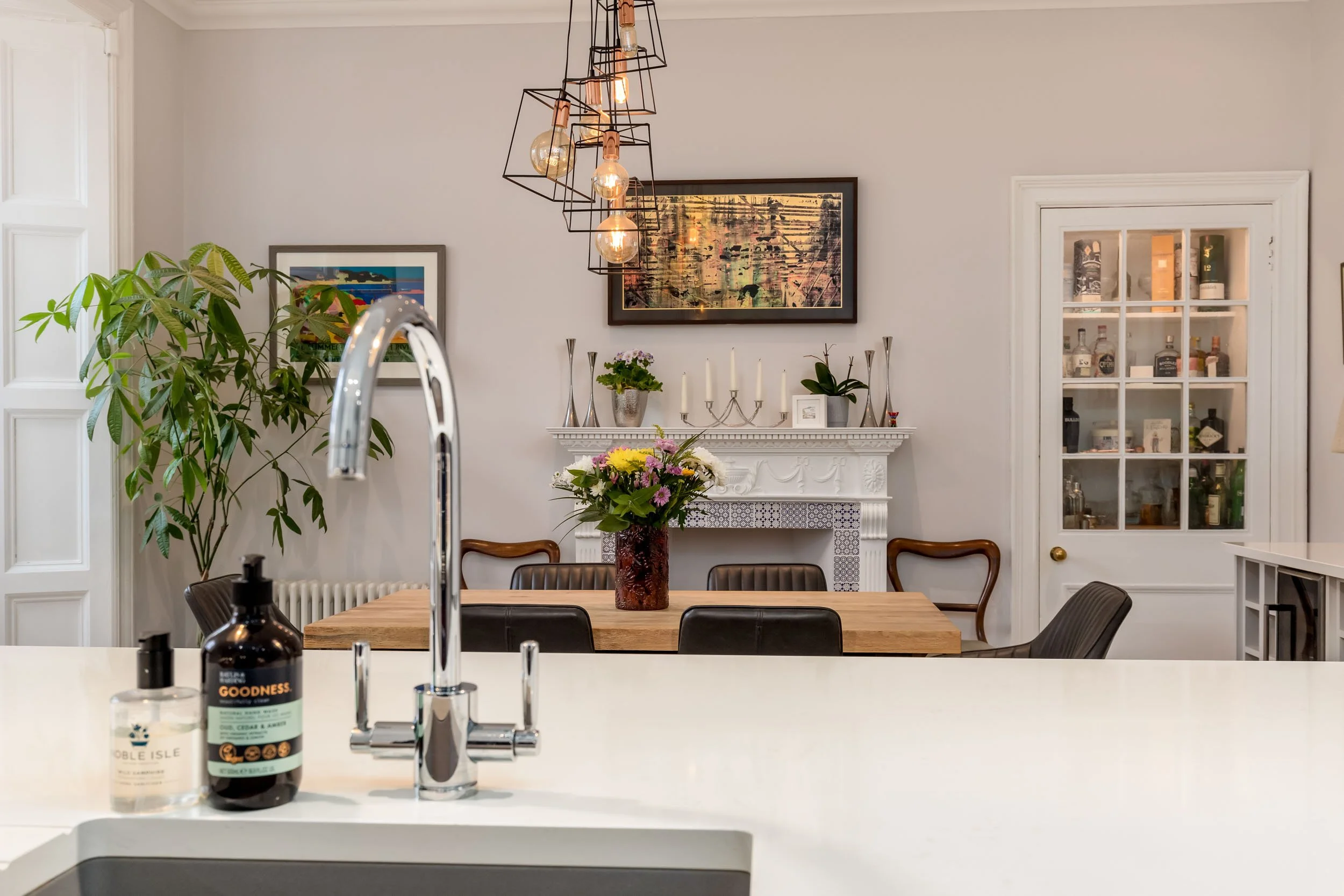 View of a dining room with a wooden table, flowers in a vase, a fireplace with decorative items, framed artwork, a glass-fronted cabinet, and a hanging light fixture.
