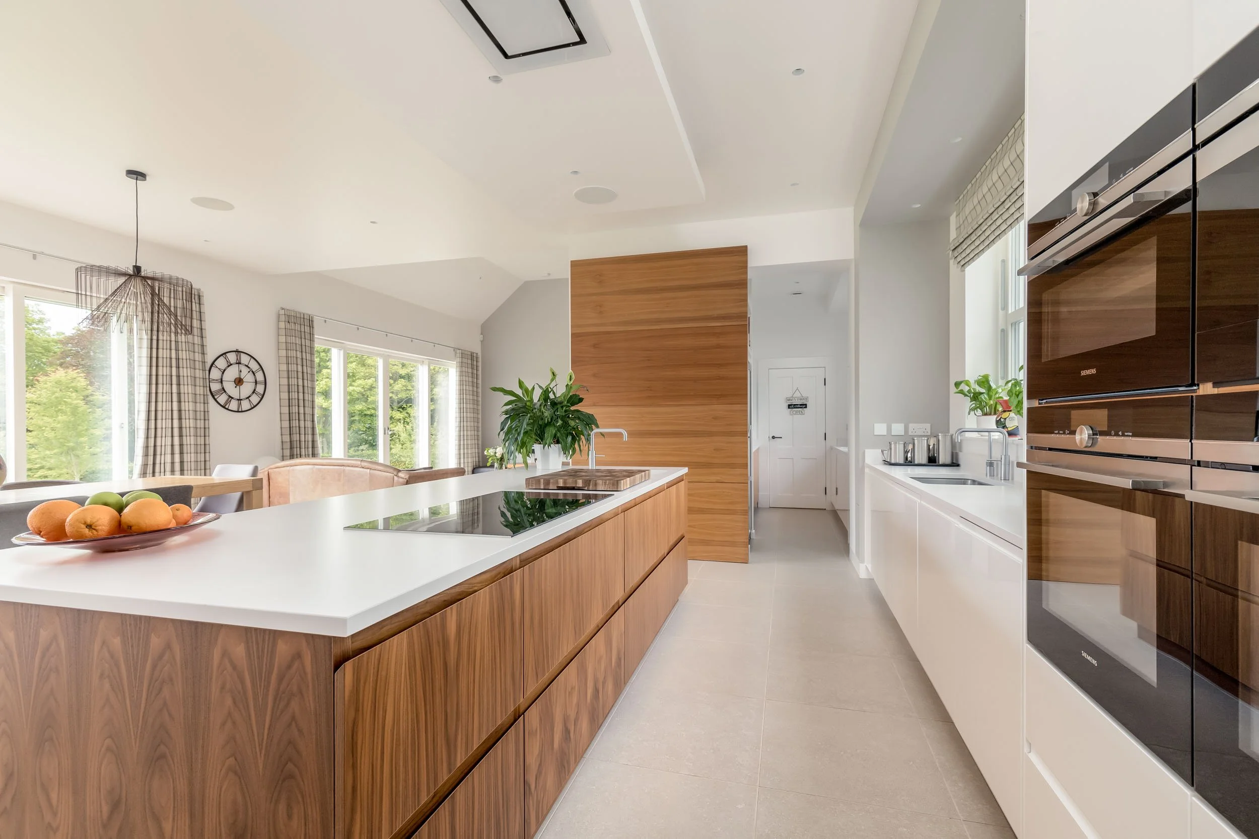 Modern kitchen with white and wooden cabinets, a large white island with a cooktop, and a view into a bright living area with large windows and checkered curtains.