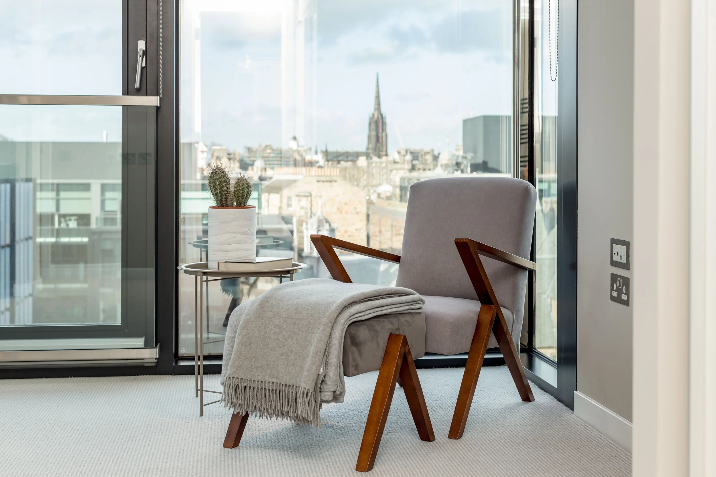 A cozy corner with a beige armchair next to a small side table holding a potted cactus and a book, in front of large glass windows showing a city skyline with a church steeple in the background.