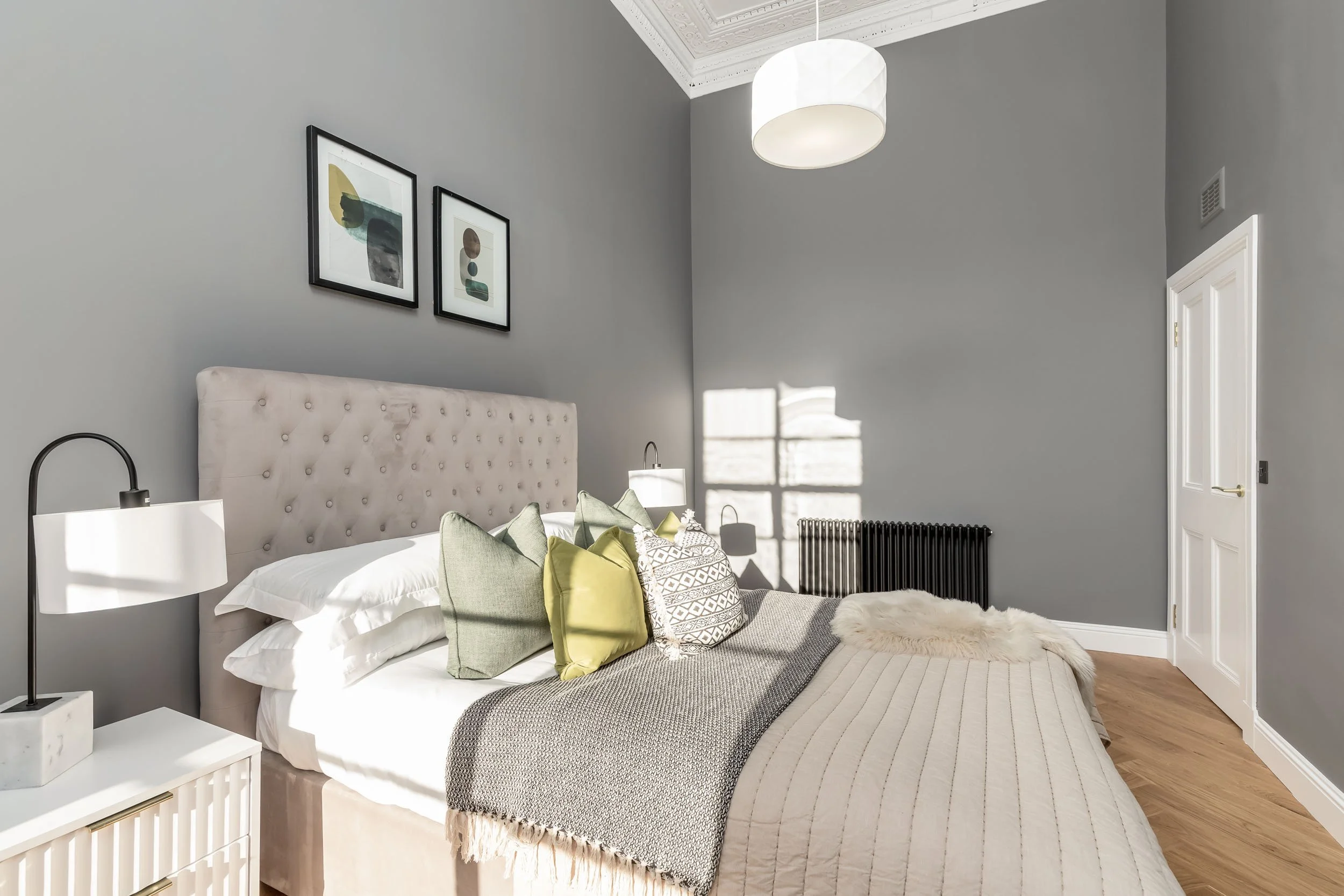 A cozy bedroom with a tufted beige headboard, gray walls, and abstract art prints. The bed has white sheets, green and patterned pillows, and a beige blanket. A fluffy white throw is at the foot. Two black wall-mounted lamps flank the bed. A white be