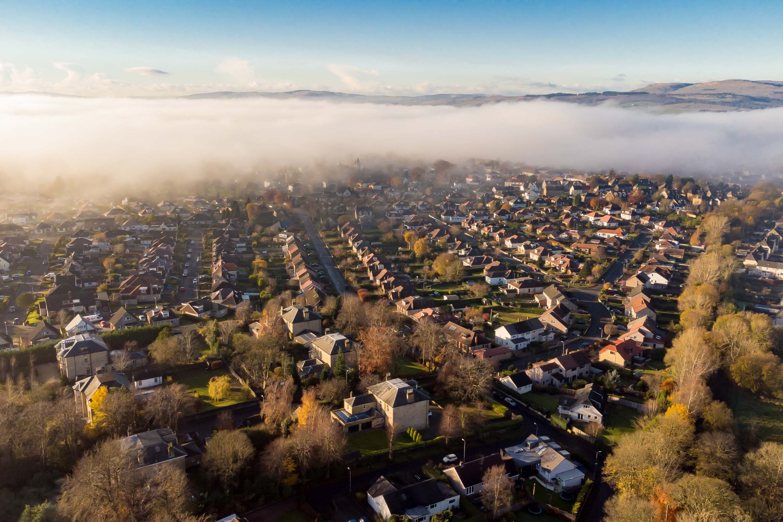 Aerial view of a residential neighborhood in the morning with fog covering the background hills and parts of the town, showing houses, trees, and streets.