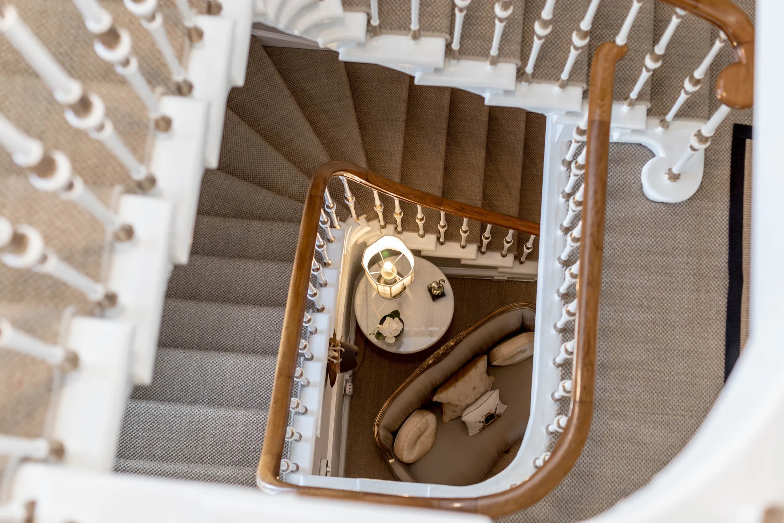 Overhead view of a spiral staircase with beige carpet, wooden handrails, and ornate white balusters, looking down onto a sitting area with a small round table, a lamp, a flower vase, and a curved sofa.