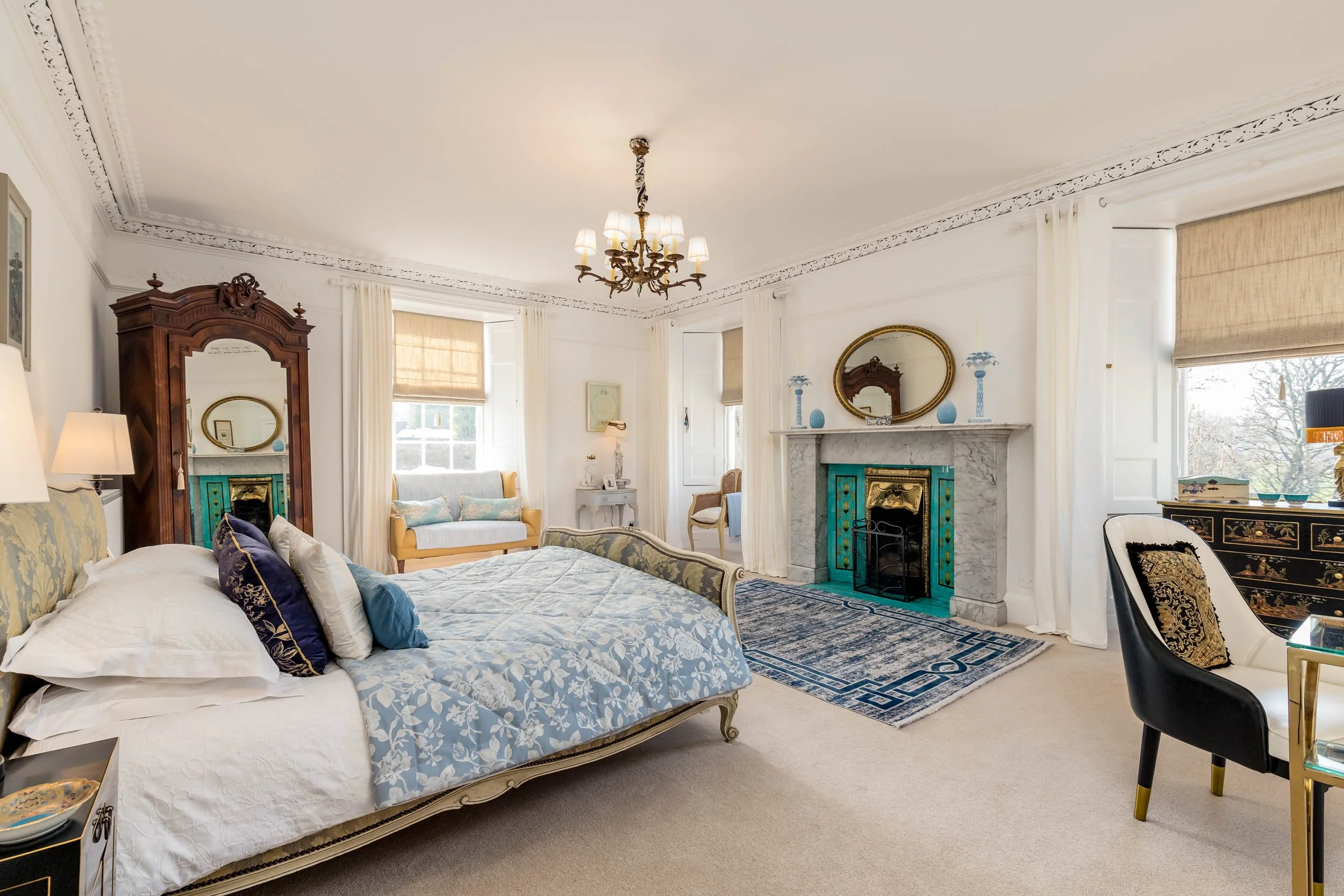 A bedroom with a Victorian-style bed, ornate wooden wardrobe, and a white marble fireplace with turquoise accents. The room features a chandelier, large windows with beige blinds, and various vintage furniture pieces and decorative pillows.