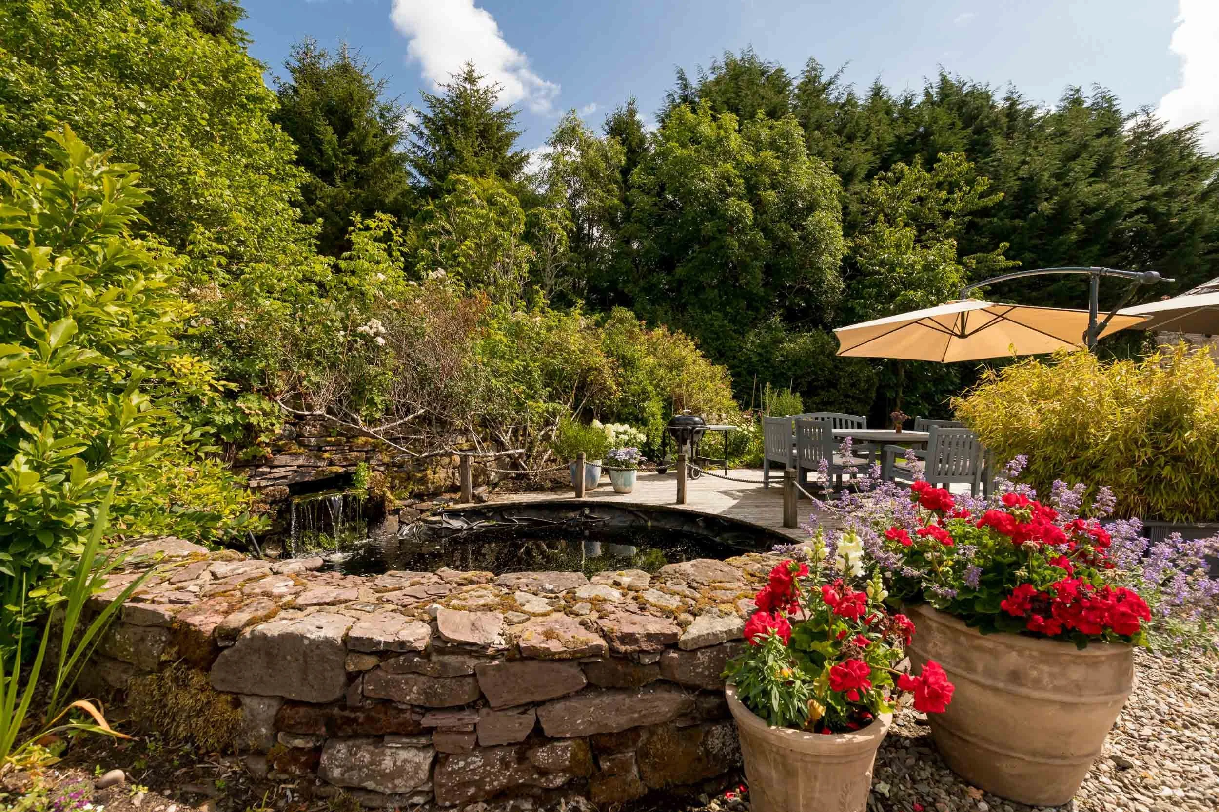 A garden patio with potted flowers, a stone water feature, outdoor dining table with umbrella, surrounded by lush green trees and plants under a partly cloudy sky.