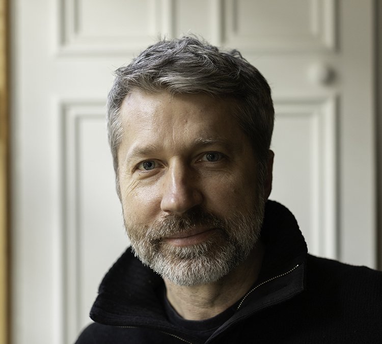 Close-up of a middle-aged man with gray hair and a beard, wearing a black zip-up jacket, standing indoors near a white door.