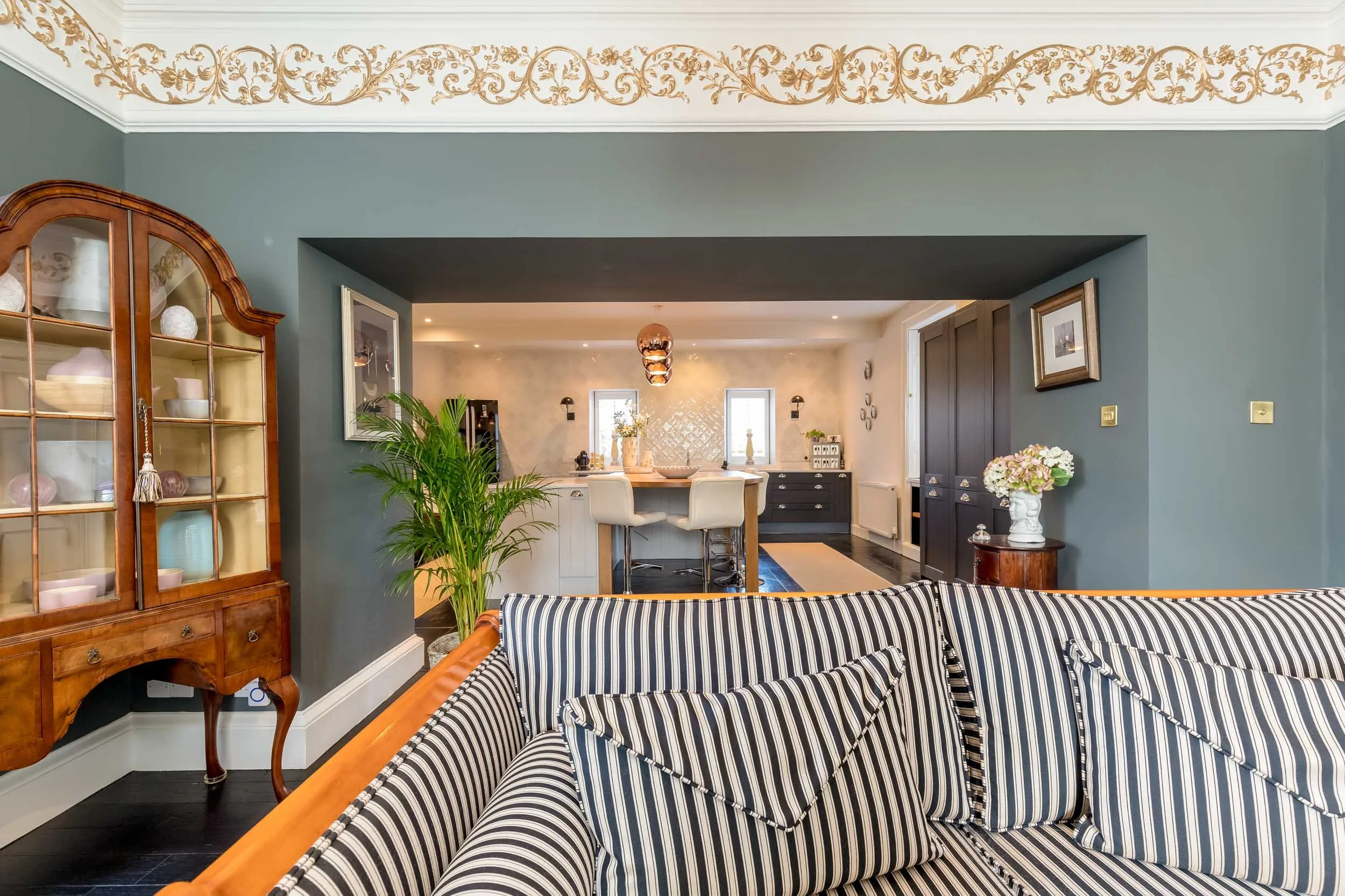 Living room with striped sofa, wooden display cabinet, and view into open kitchen with barstools.