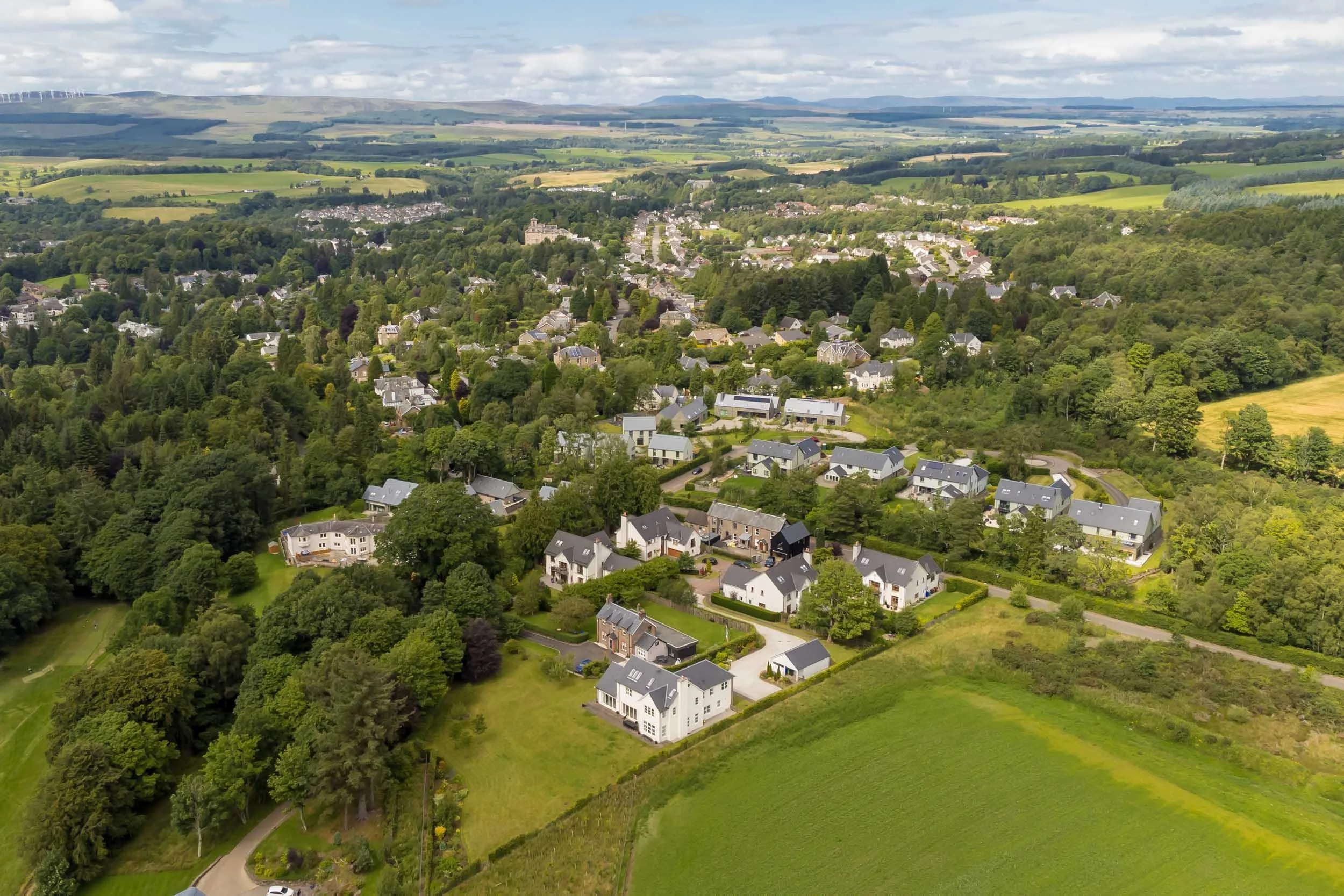 Aerial view of a suburban neighborhood with houses surrounded by green trees and fields, under a partly cloudy sky.