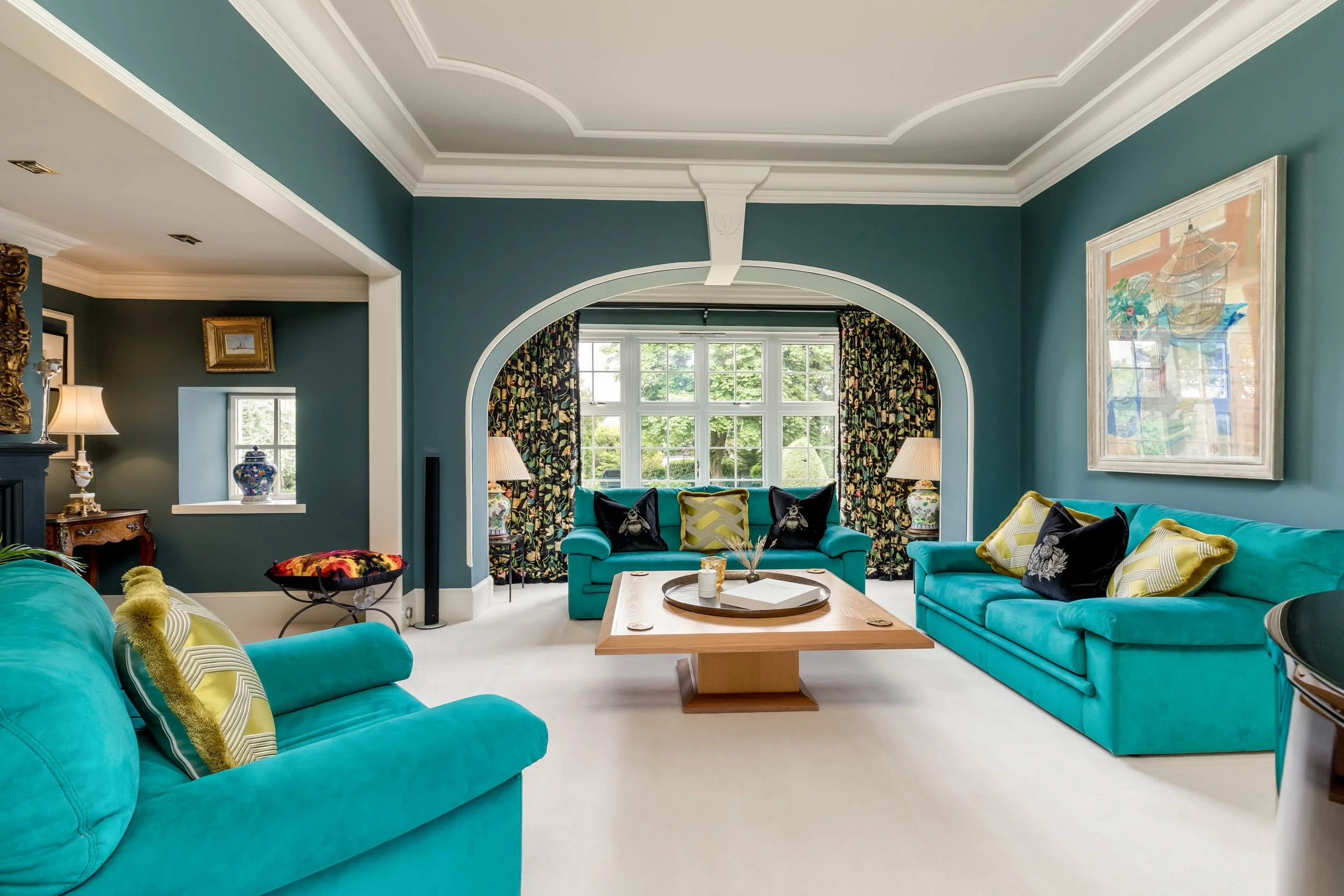 Living room with three turquoise sofas, decorative pillows, and a large window with floral curtains, hardwood coffee table, and art on the walls.