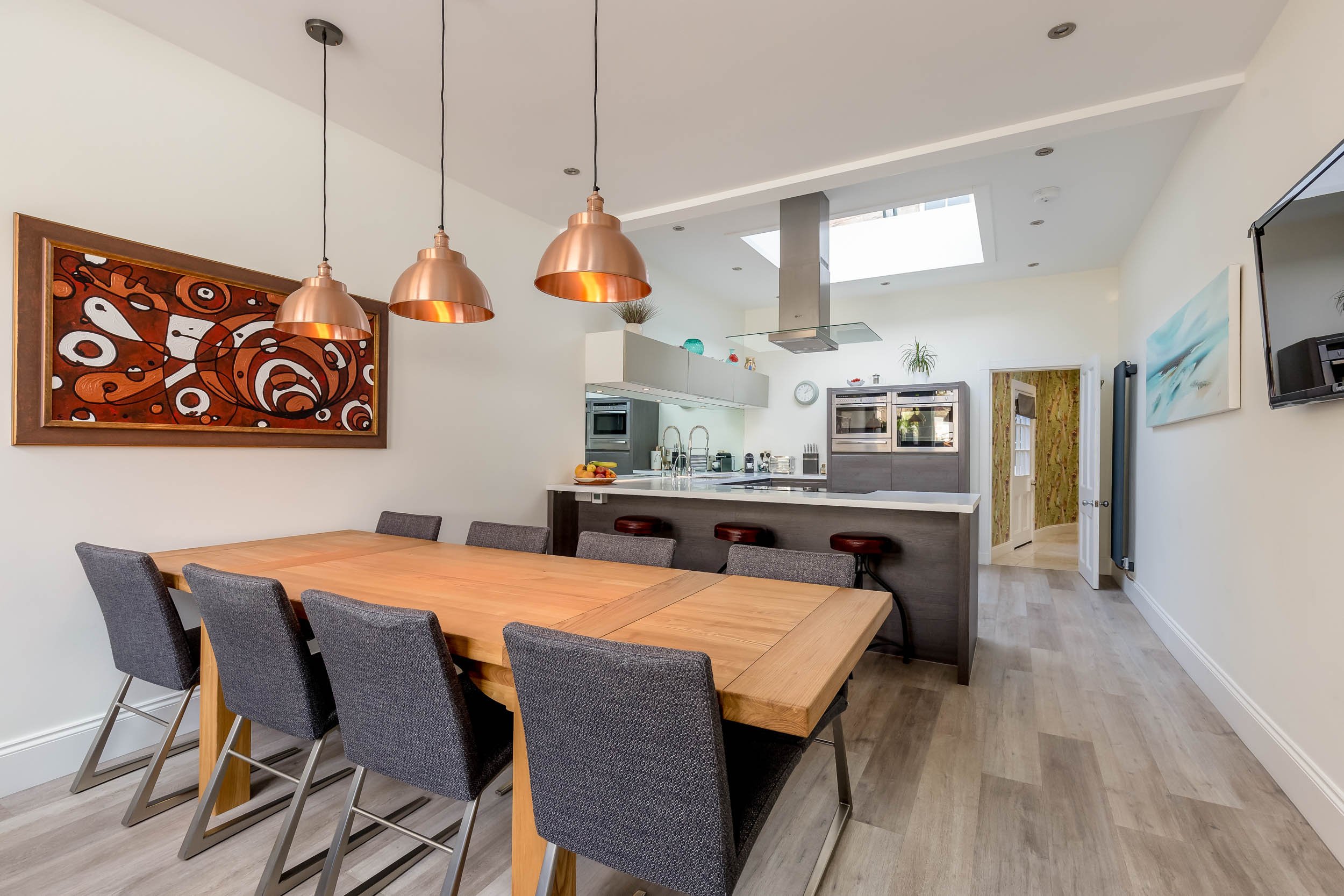 A modern open-concept kitchen and dining area with a wooden dining table, gray chairs, and three copper pendant lights. The kitchen has a white countertop, gray cabinets, and built-in appliances. A colorful abstract painting hangs on the wall, and a 