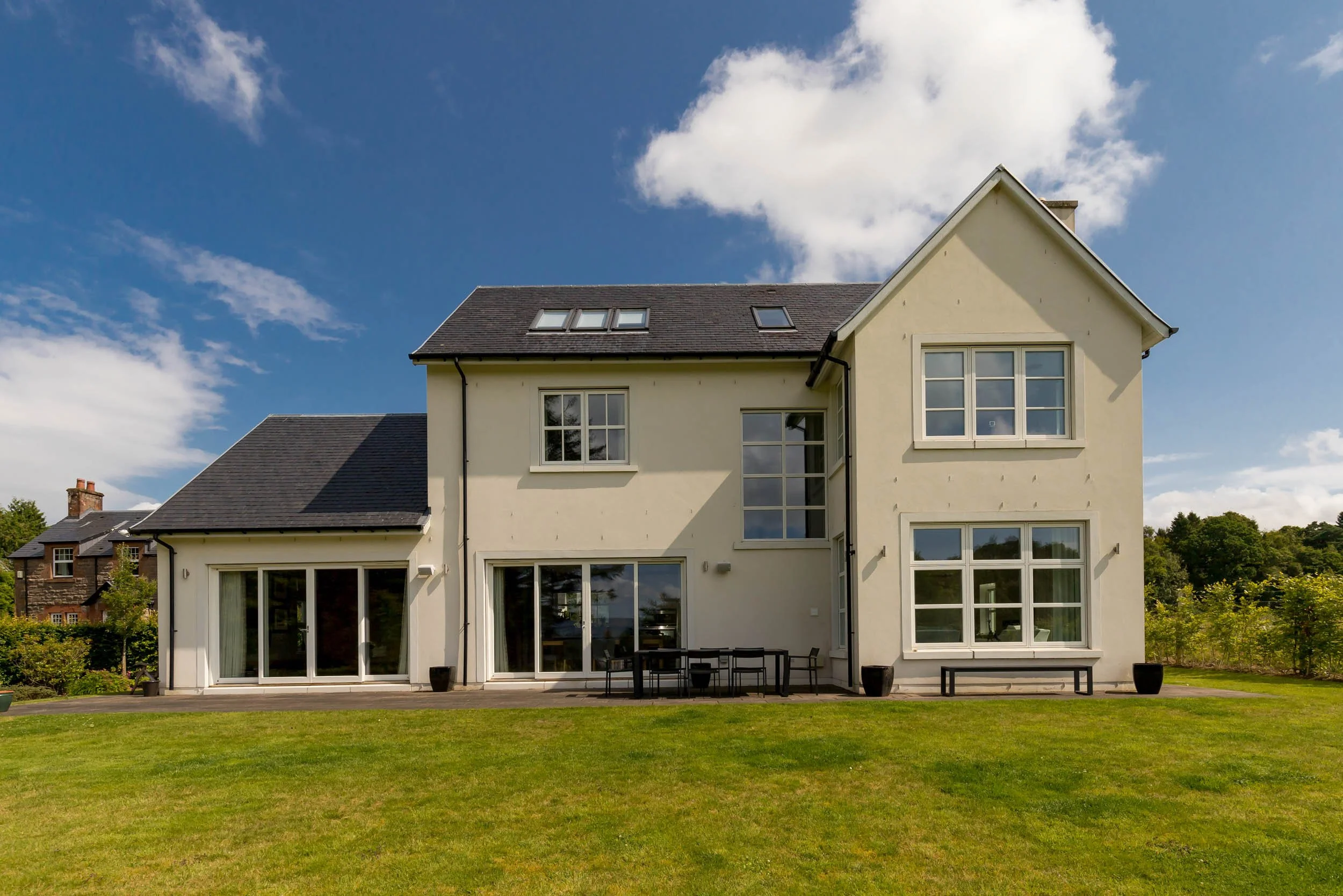 A white, multi-story house with large windows, set against a bright blue sky with clouds, and surrounded by a green lawn.