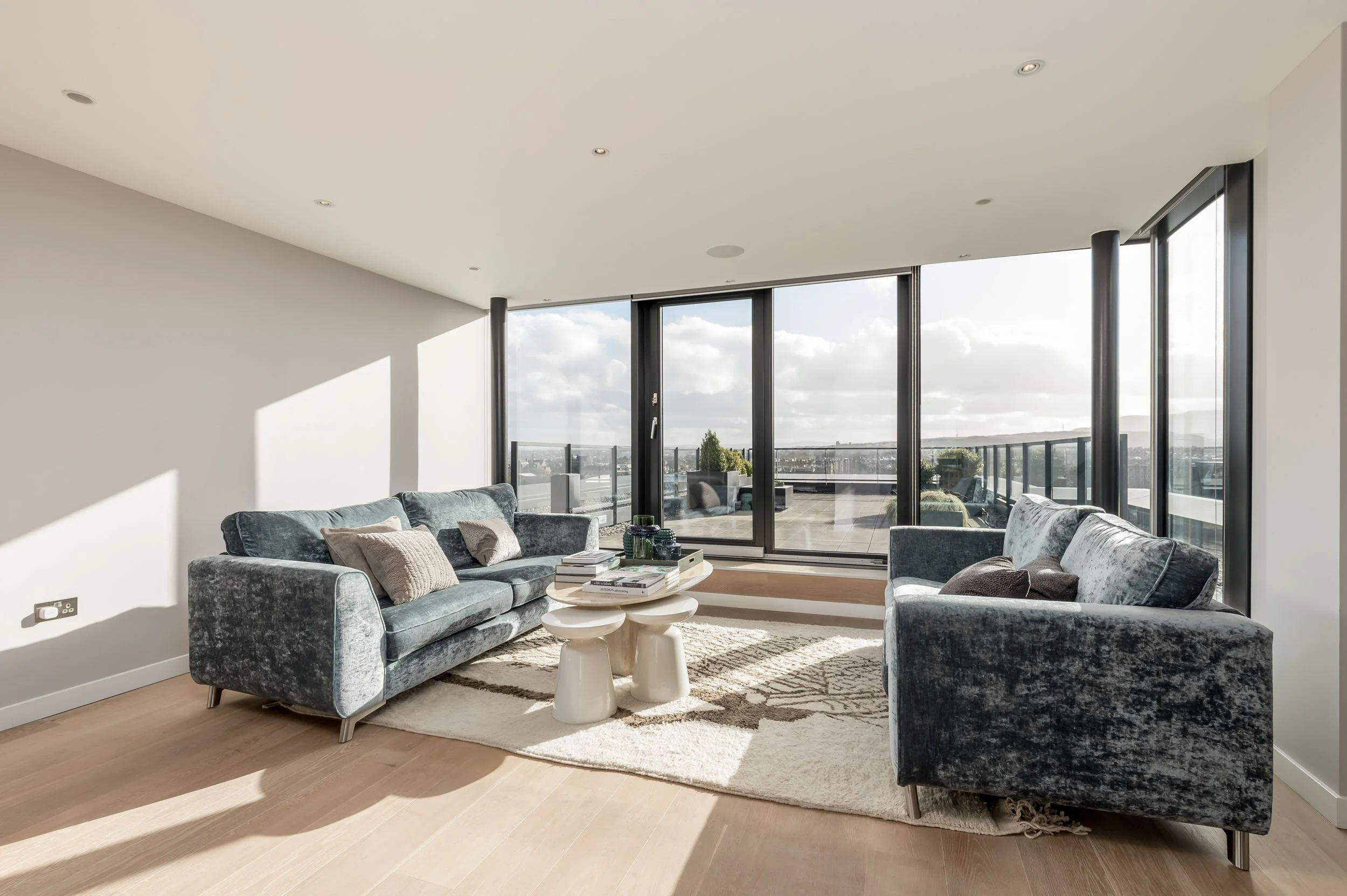 Modern living room with large glass sliding doors leading to a balcony, two velvet sofas with cushions, a round coffee table with books and decor, light wood flooring, and natural sunlight streaming in.