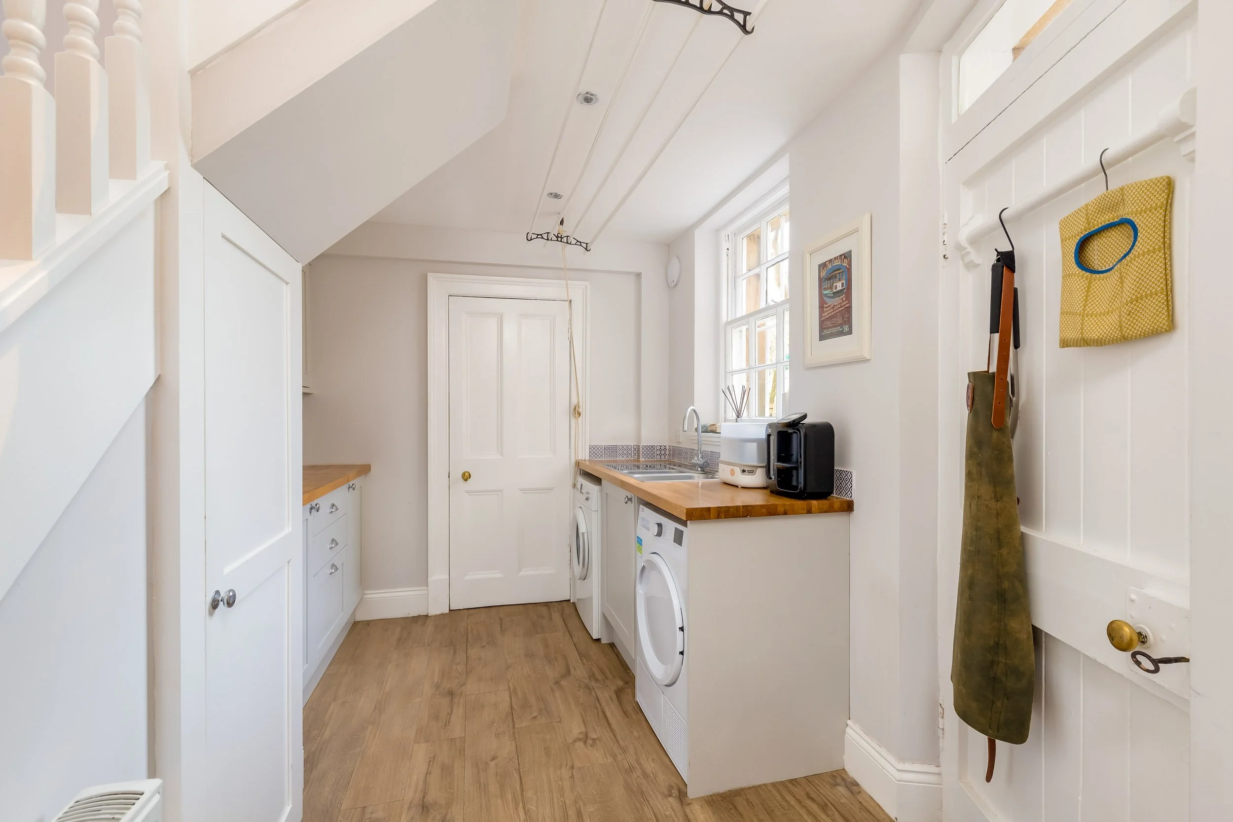 A laundry room with white walls, wooden floors, a window, and white cabinets. There are washing machine and dryer beneath a wooden countertop, with various appliances on top. A door with a hook holds an apron, and a framed picture hangs on the wall.