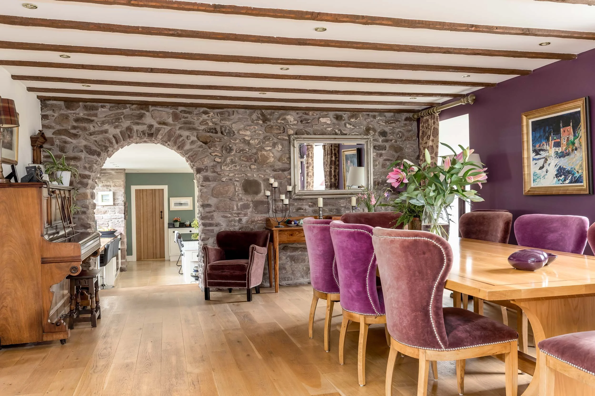 A living room and dining area with a stone archway, wooden beams on the ceiling, purple and brown velvet chairs, a wooden dining table decorated with a vase of pink lilies, a piano, and framed artworks on the walls.
