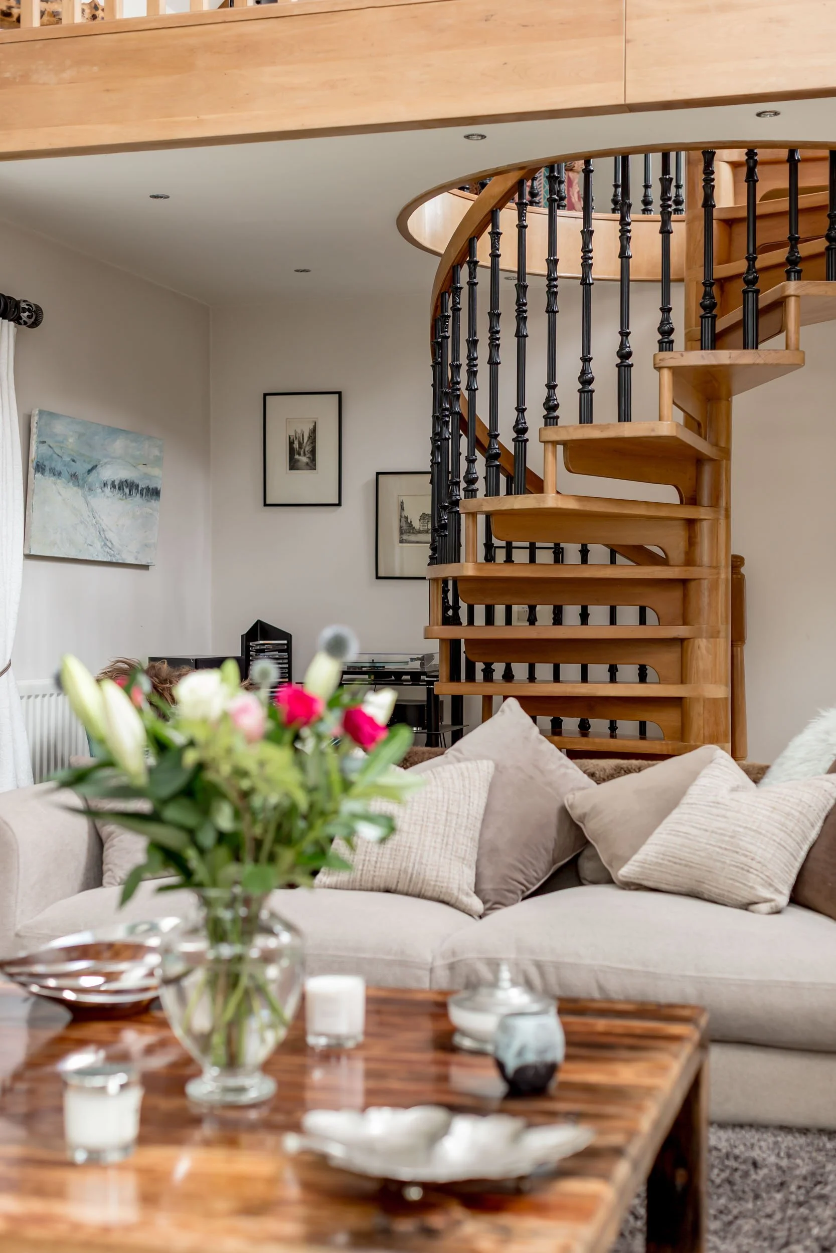Living room with beige sofa and pillows, wooden spiral staircase with black balusters, wall art, and a coffee table with a vase of flowers.