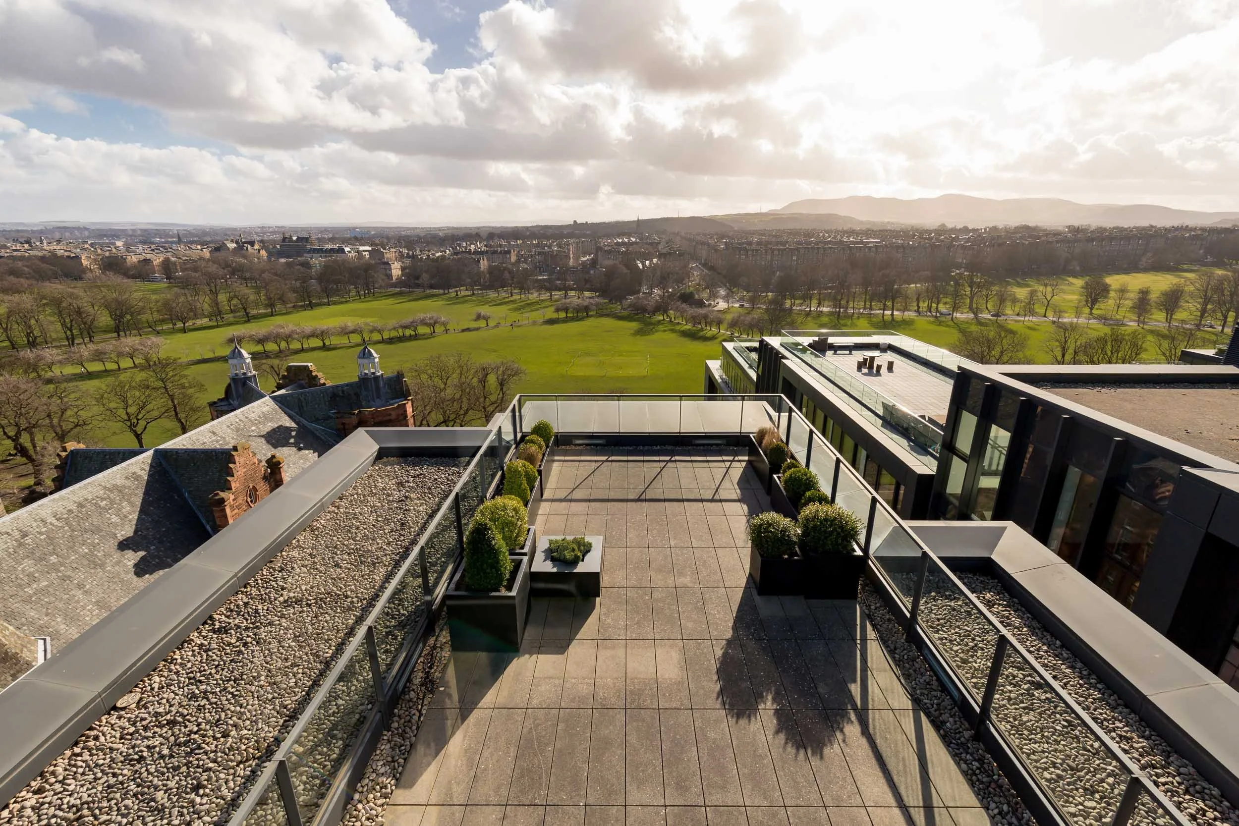 A rooftop terrace with potted bushes overlooking a city park and skyline in the distance.