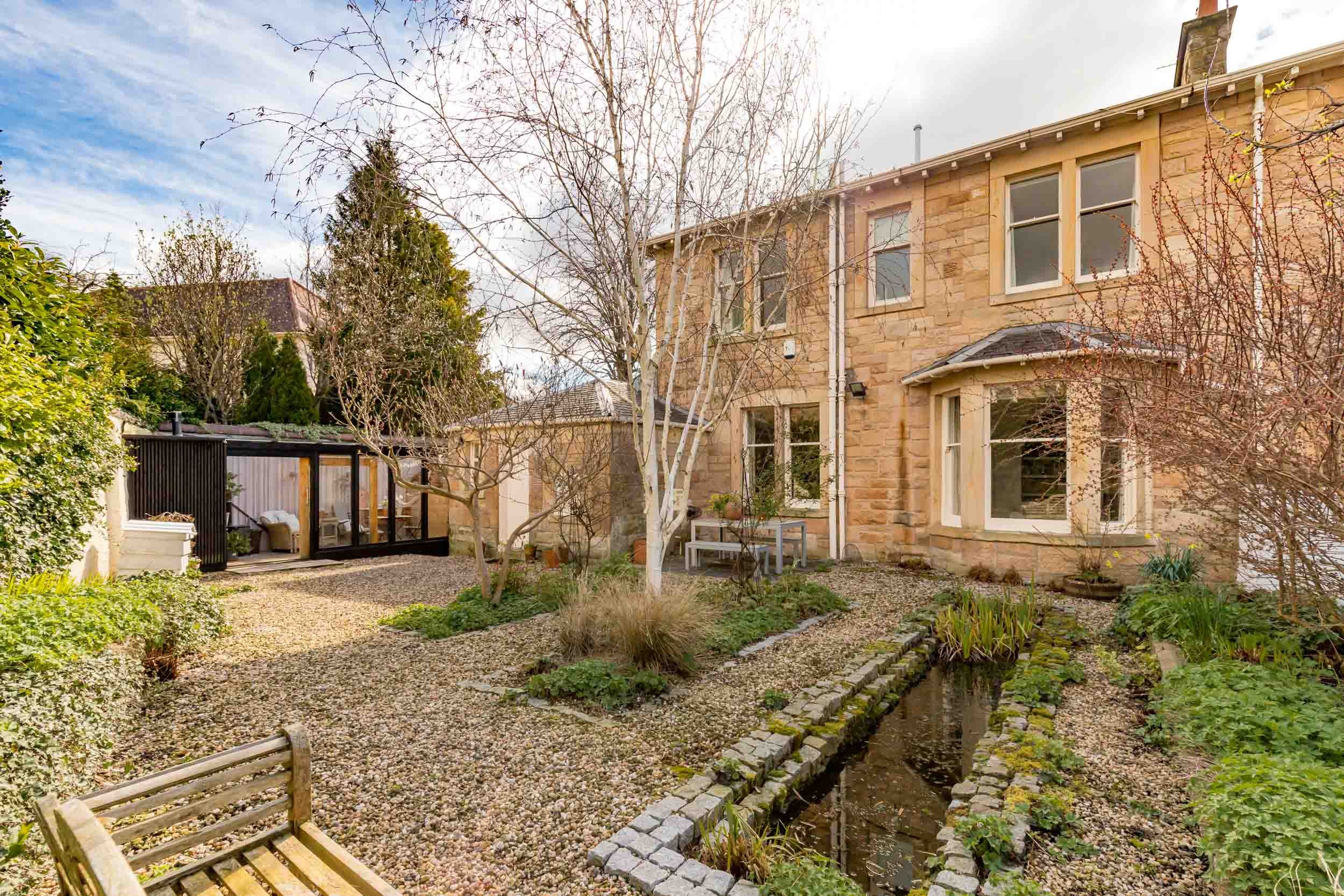 Backyard garden with leafless trees, a small pond bordered with bricks, a gravel pathway, and a stone house with multiple windows.
