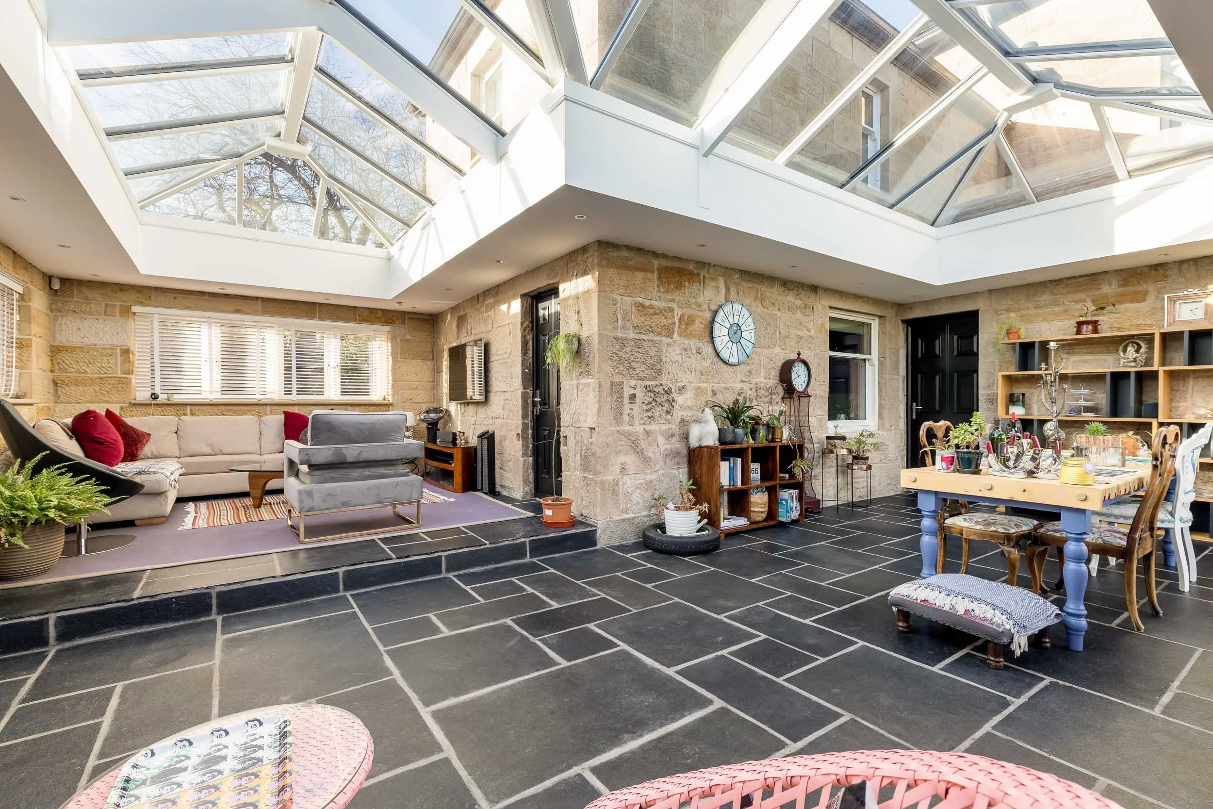 Sunroom with large skylight, stone walls, black tile flooring, beige sectional sofa with red pillows, wooden shelving unit with plants, dining table with chairs, and various decorative items.