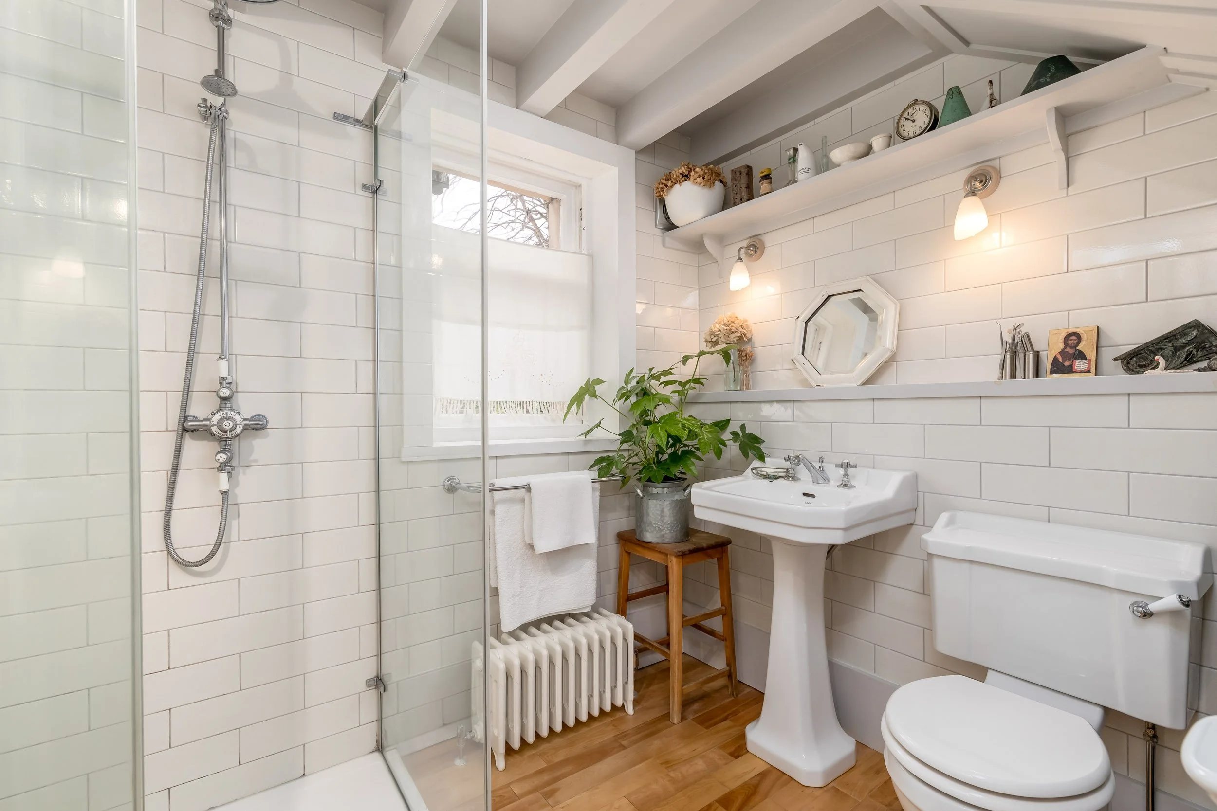 Bathroom with a walk-in glass shower, white tiled walls, a sink with a pedestal, a toilet, a window with a sheer curtain, a plant on a small wooden stand, and decorative items on a shelf.