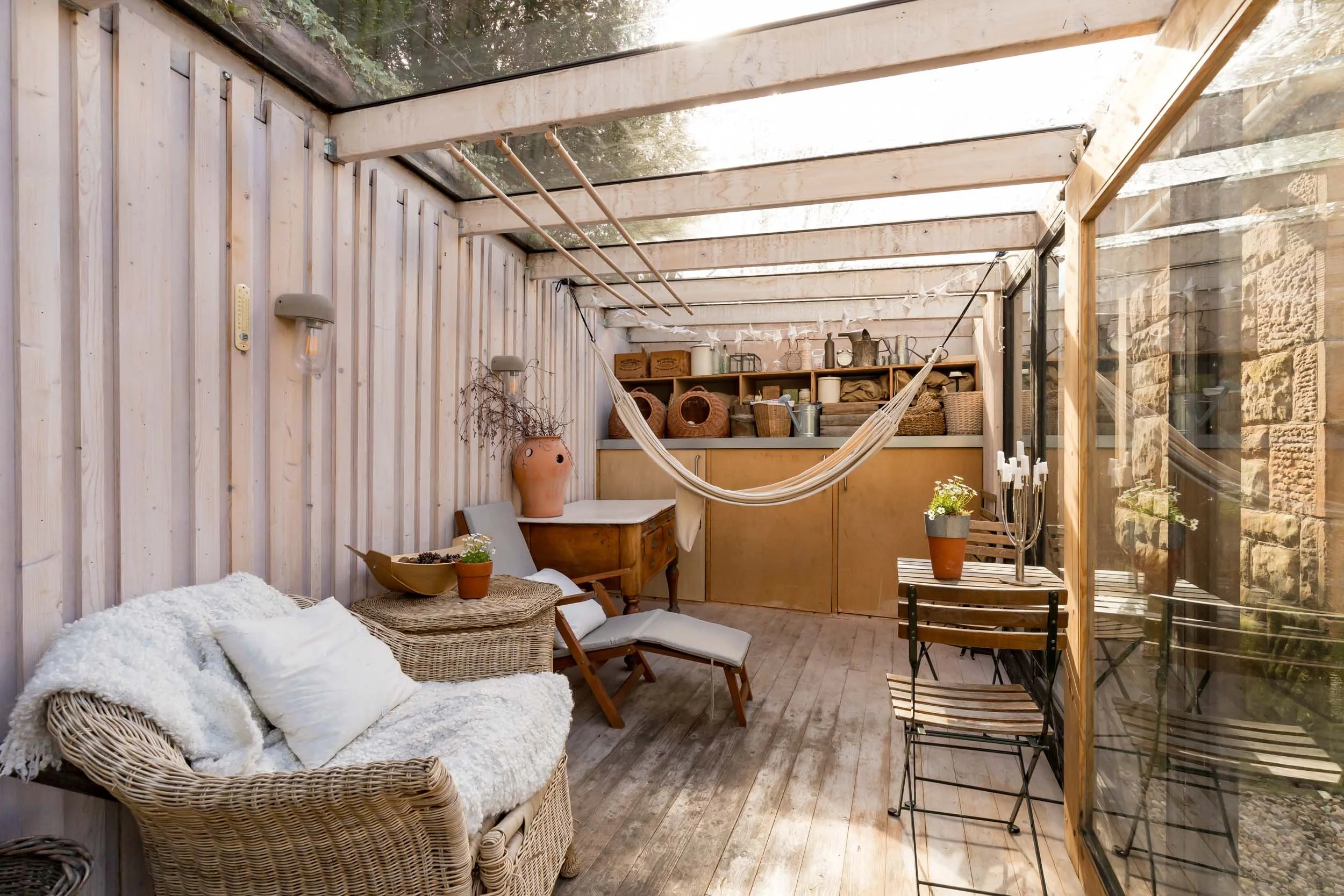 Cozy enclosed patio with wicker sofa, wooden chairs, hammock, and shelves with decorative items and plants.