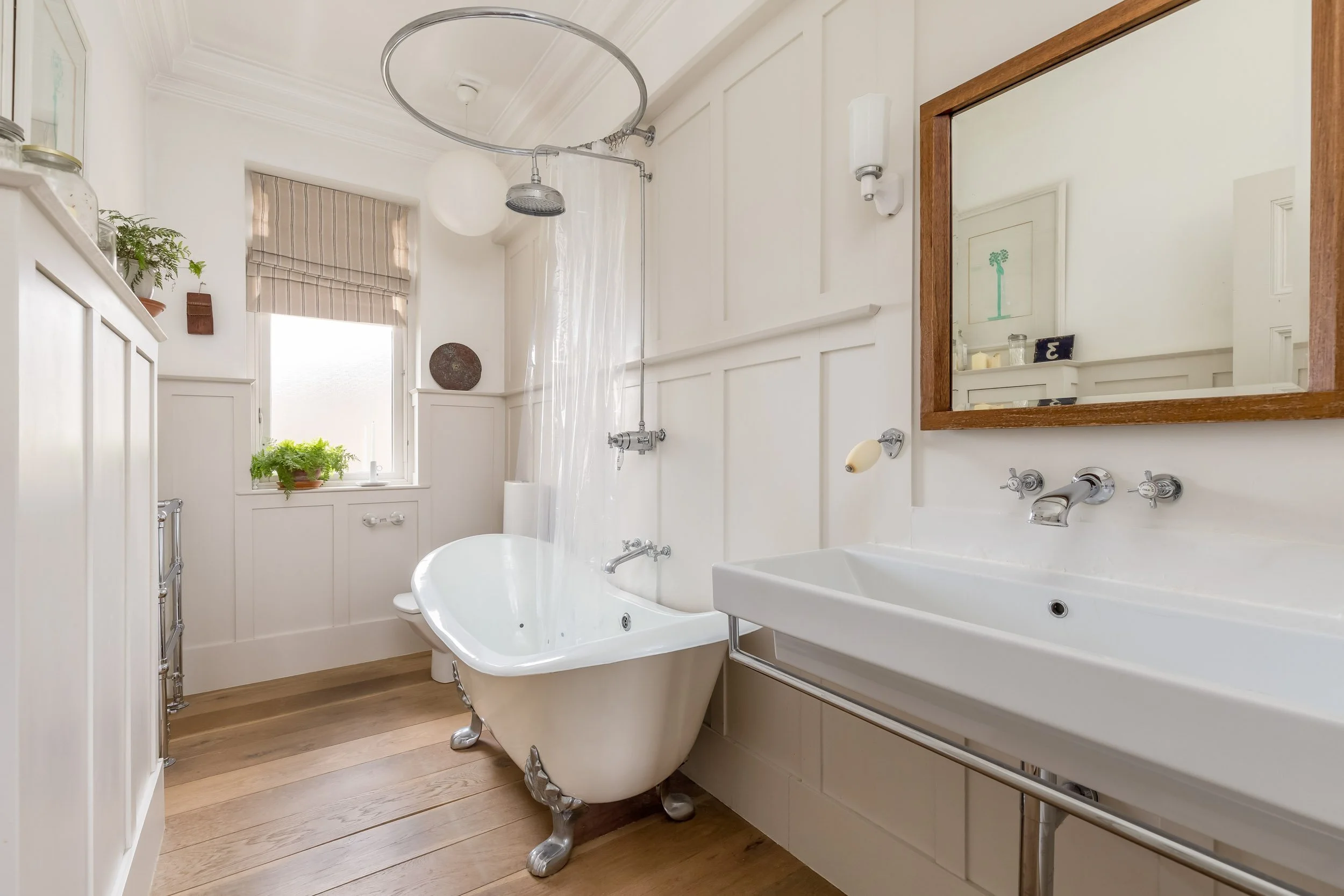 A bright bathroom with a freestanding clawfoot bathtub, a white sink with a mirror above, wooden flooring, and a window with potted plants on the windowsill.