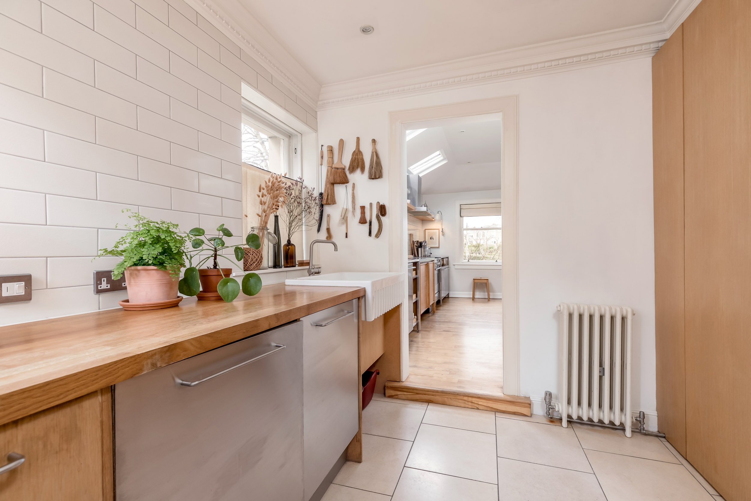 A kitchen with white tiled walls, wooden countertop, potted plants, window, and a doorway leading to a bright dining area with wooden floors and large windows.