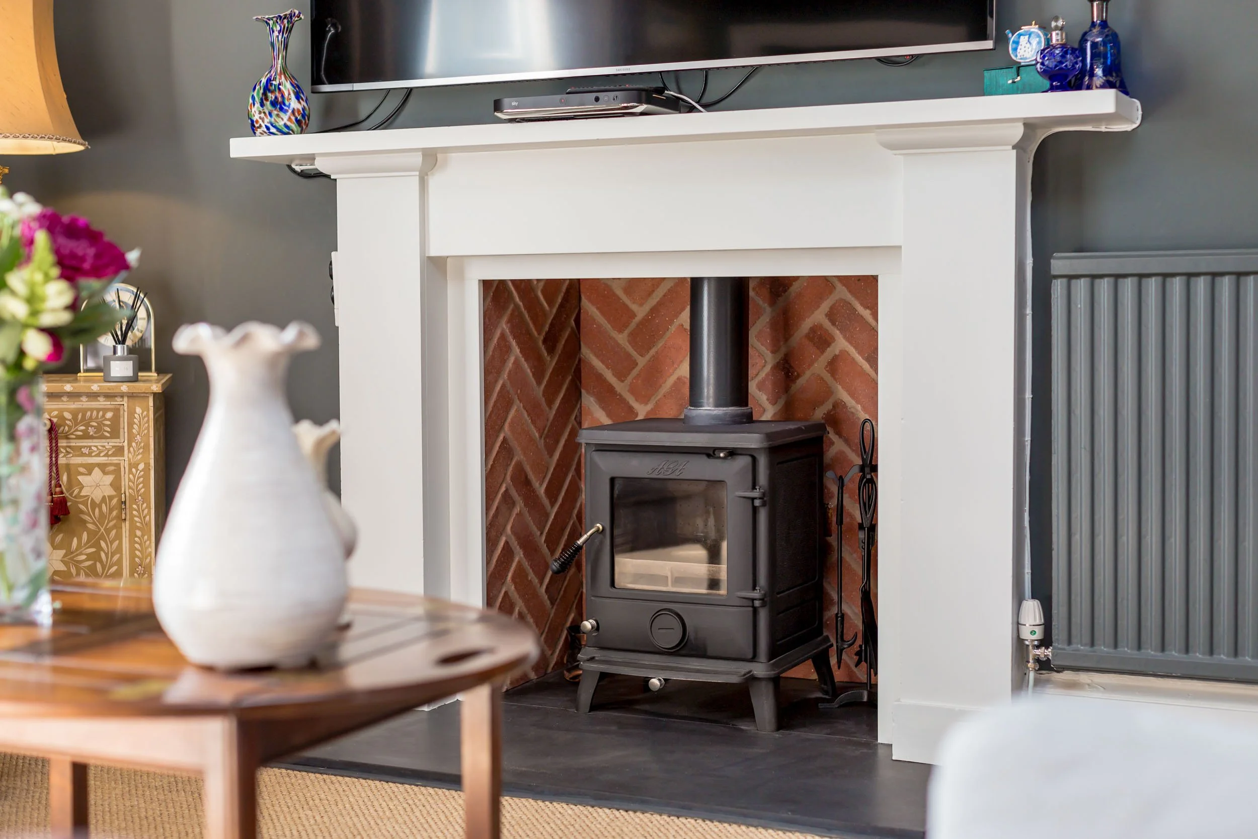 Living room fireplace with a black wood stove, white mantel, and a brick interior. A flat-screen TV is mounted above the mantel with decorative items including a colorful vase on the left.