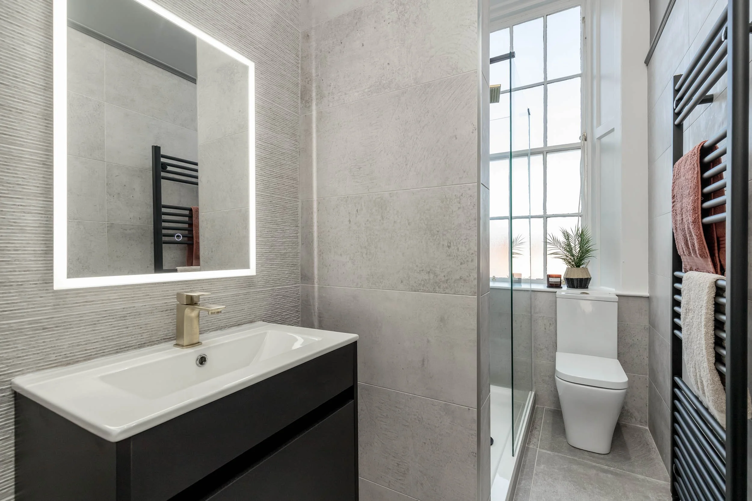 Modern bathroom with a rectangular mirror with LED lighting, a white sink with a black vanity, a walk-in glass shower, a white toilet, a large window with a potted plant on the windowsill, and a black towel radiator with folded towels.