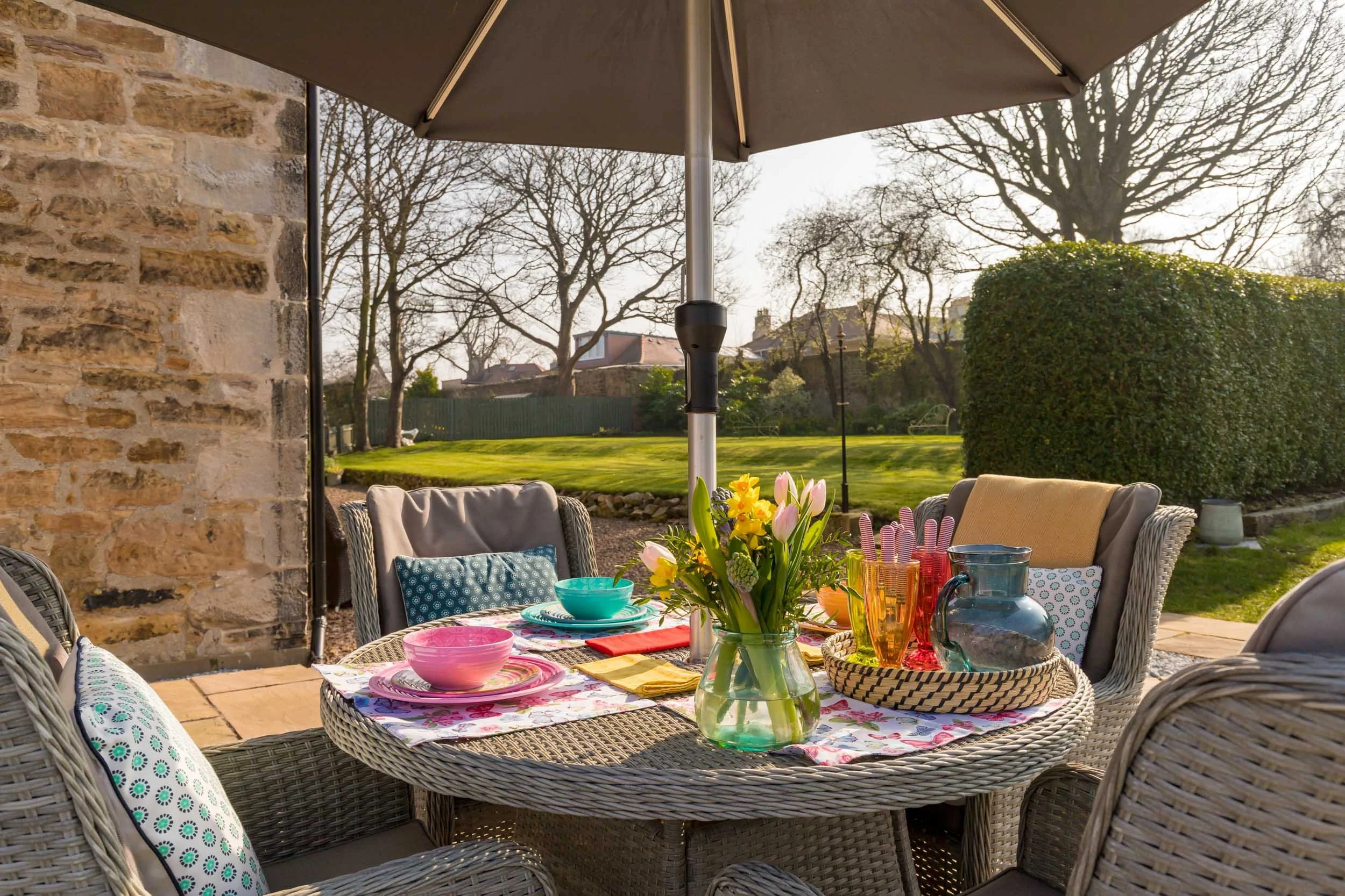 Outdoor patio dining setup with a wicker round table under a large patio umbrella, set with colorful bowls, glasses, a pitcher, and a flower arrangement, with a lush green garden and trees in the background.