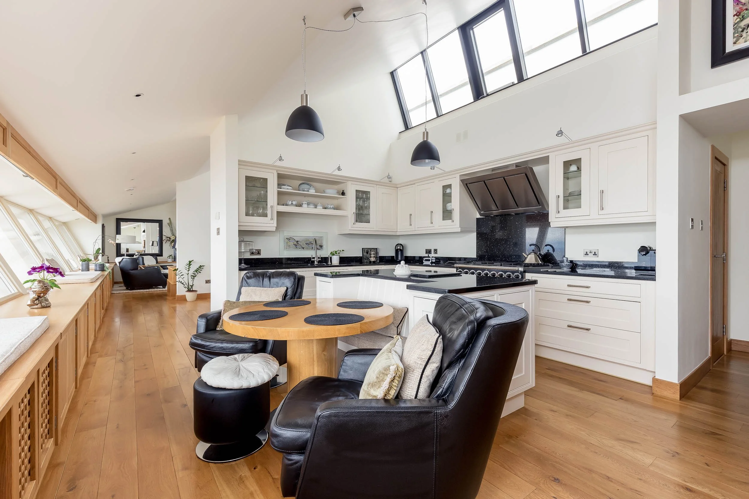 Open kitchen and dining area with white cabinets, black countertops, black appliances, wooden flooring, and large windows with skylights