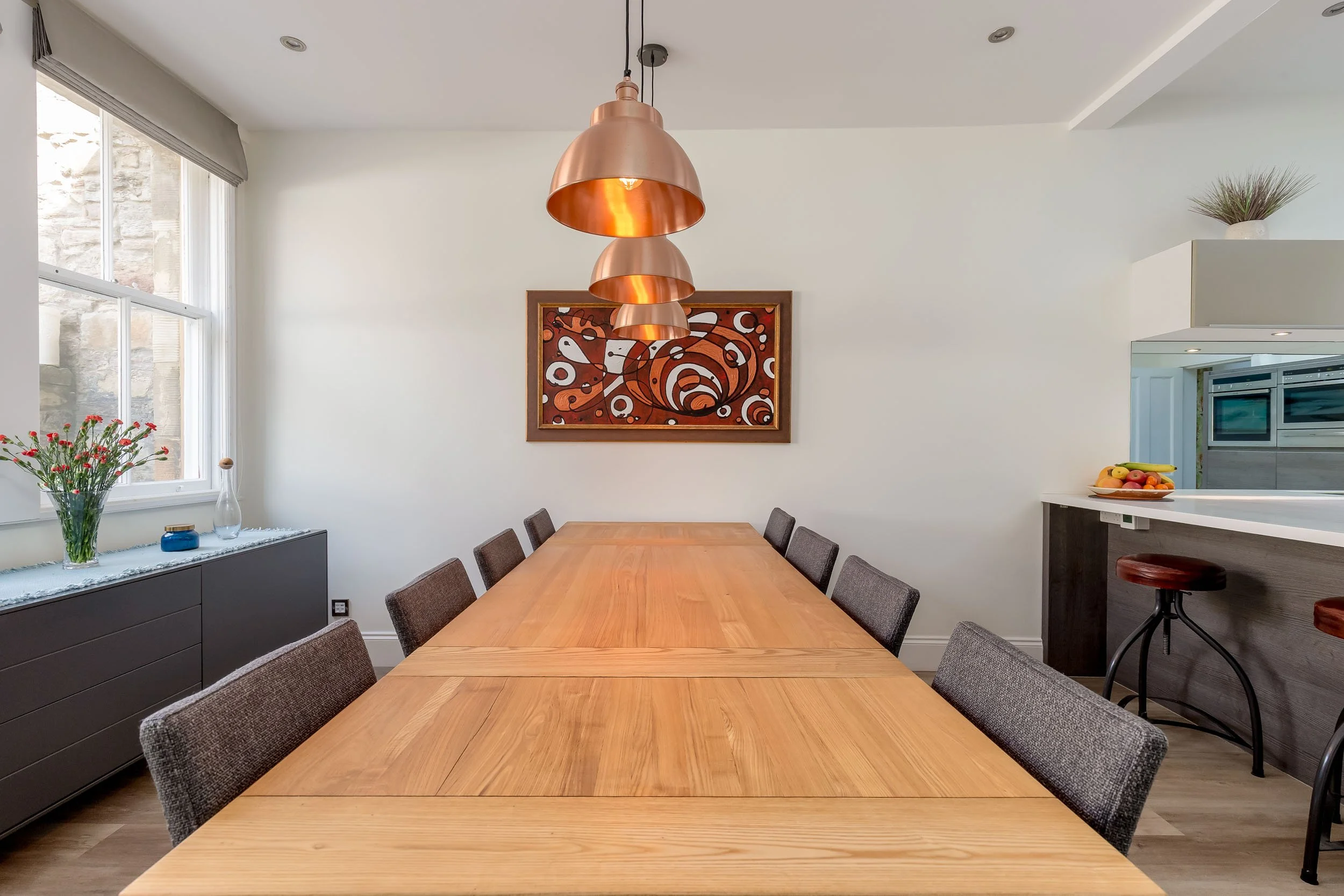 A modern dining room with a long wooden table, eight chairs, copper pendant lights, a window with a flower vase, and wall art.