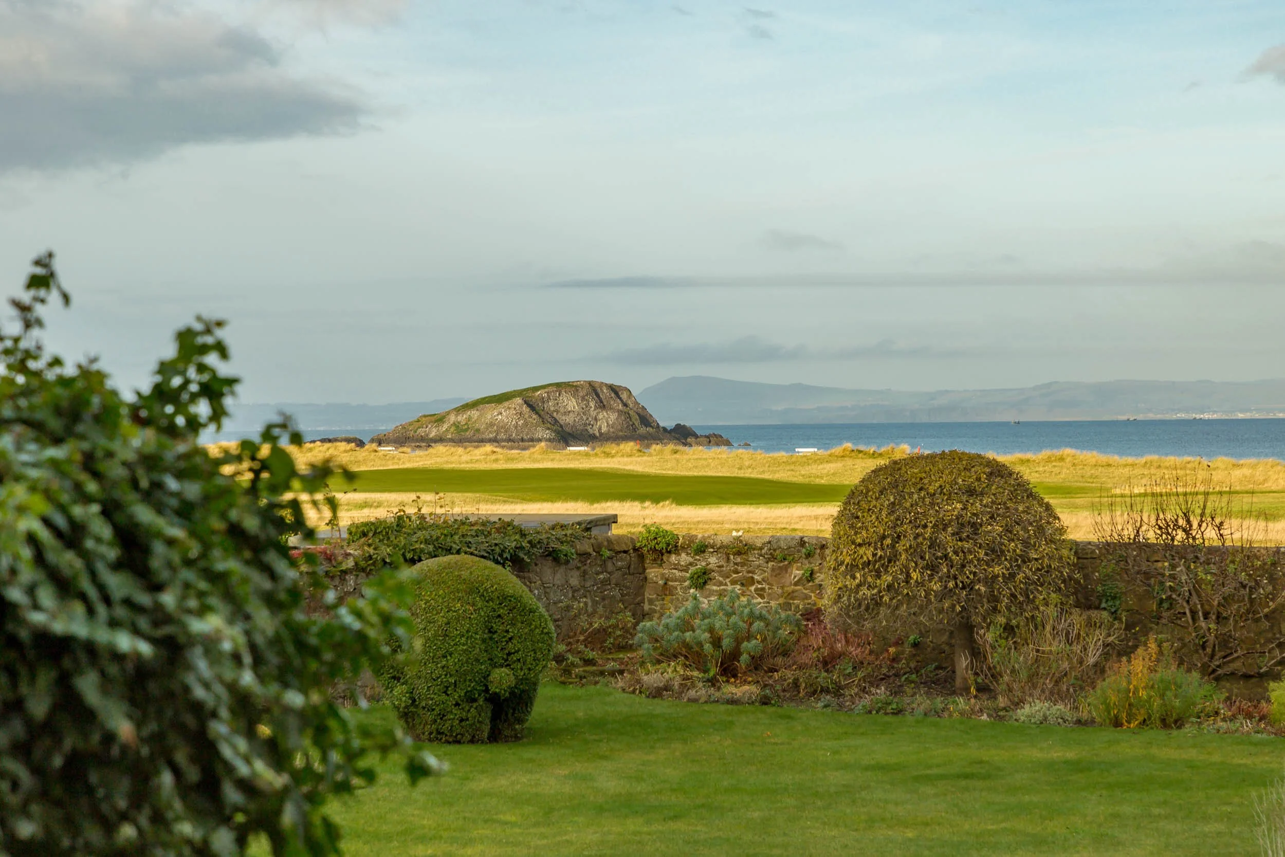 A landscaped garden with trimmed bushes and a stone wall, overlooking a grassy golf course with an island in the water and distant hills.