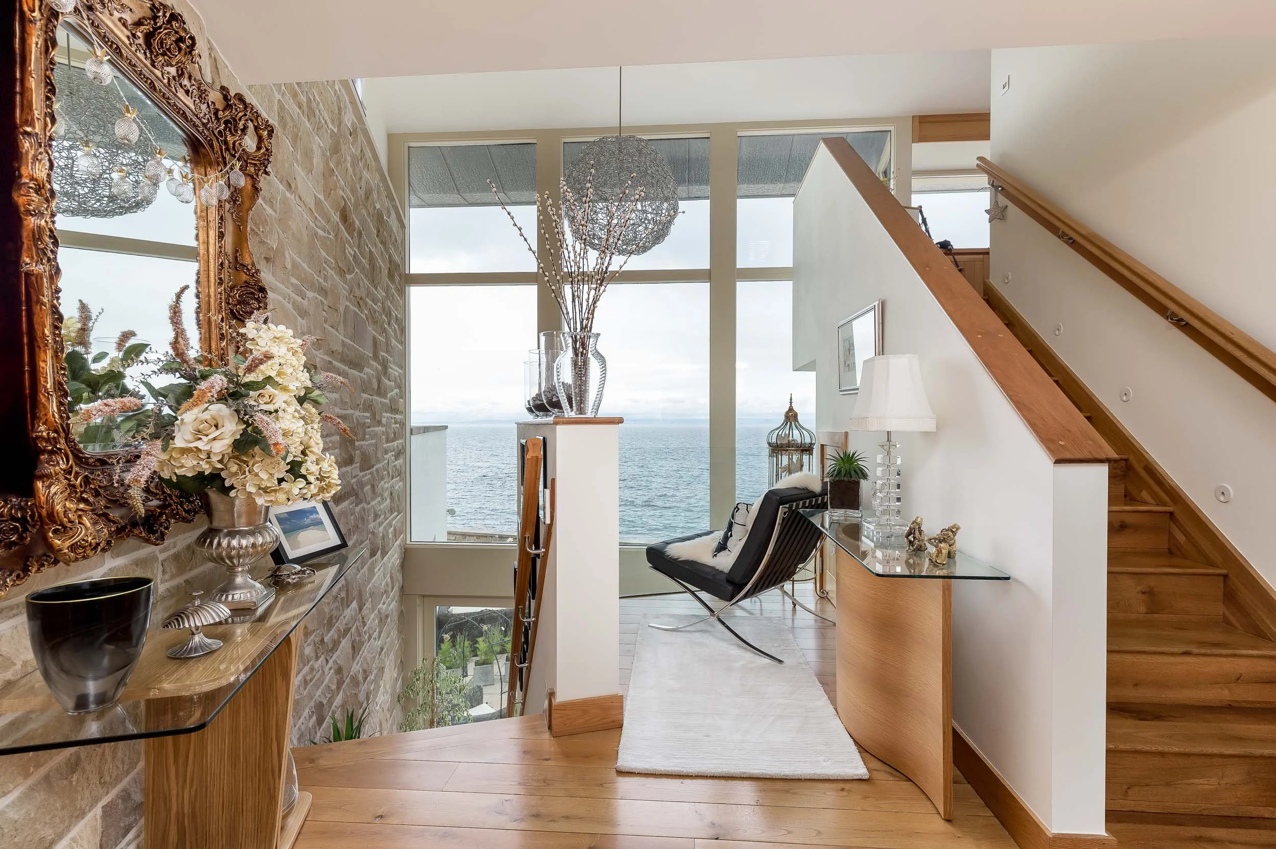 Interior of a modern living room with large windows overlooking the ocean, featuring a mix of antique and contemporary decor, a black lounge chair, and wooden accents.