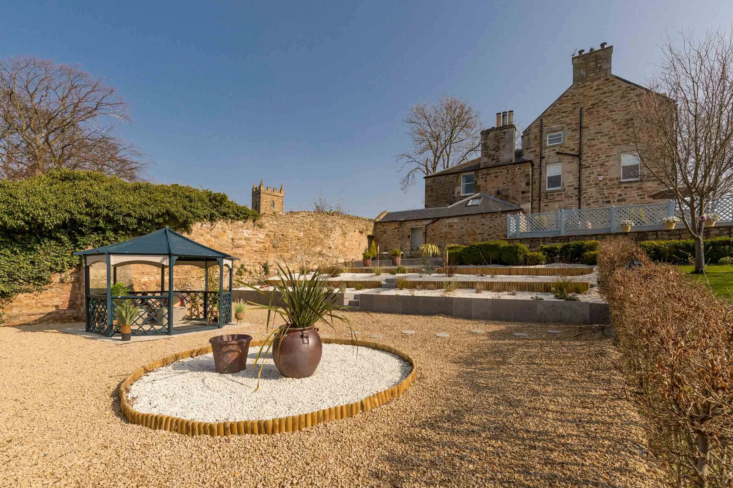 A backyard with a gravel ground, a small gazebo, potted plants, a stone house, and a stone wall with a tower in the background under a clear blue sky.