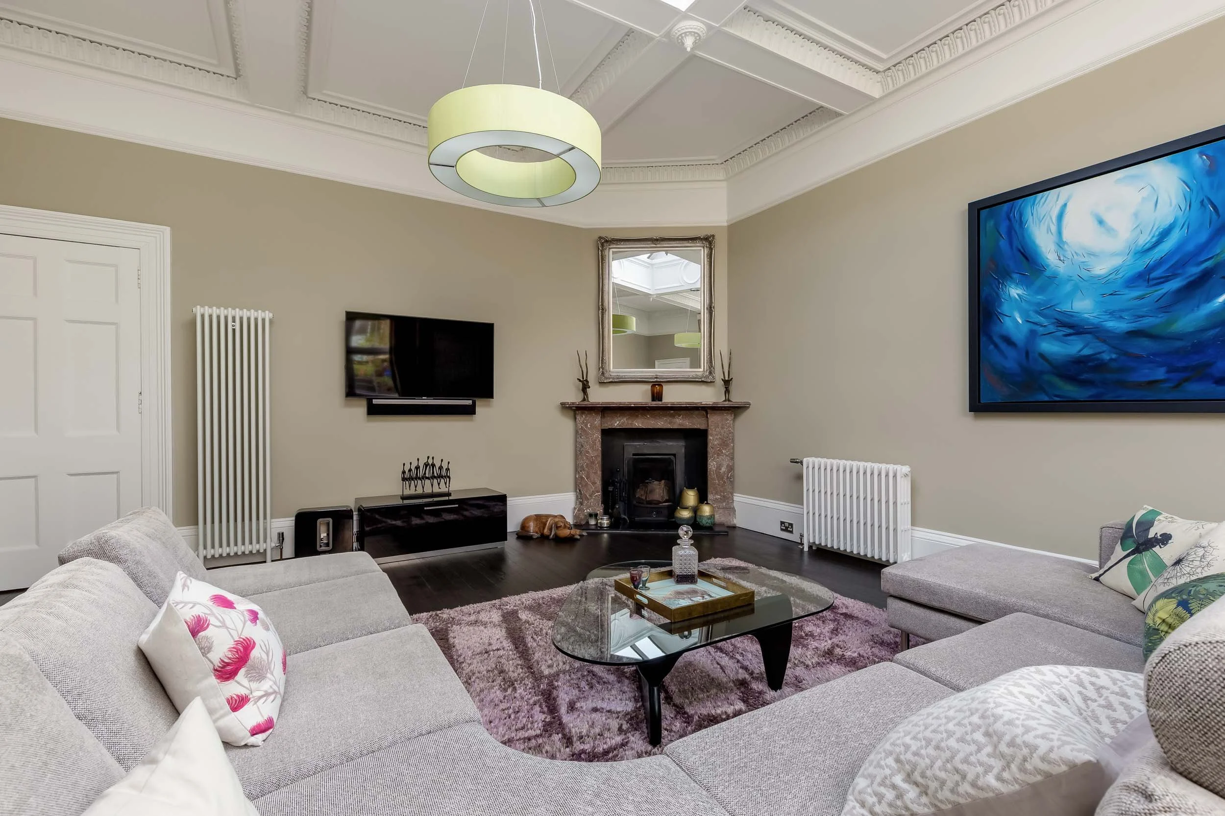 Living room with beige walls, white crown molding, and a gray sectional sofa with colorful pillows. There is a glass coffee table on a purple rug, a gray fireplace with decor, a wall-mounted TV, and a large modern blue abstract painting. A mirror is 