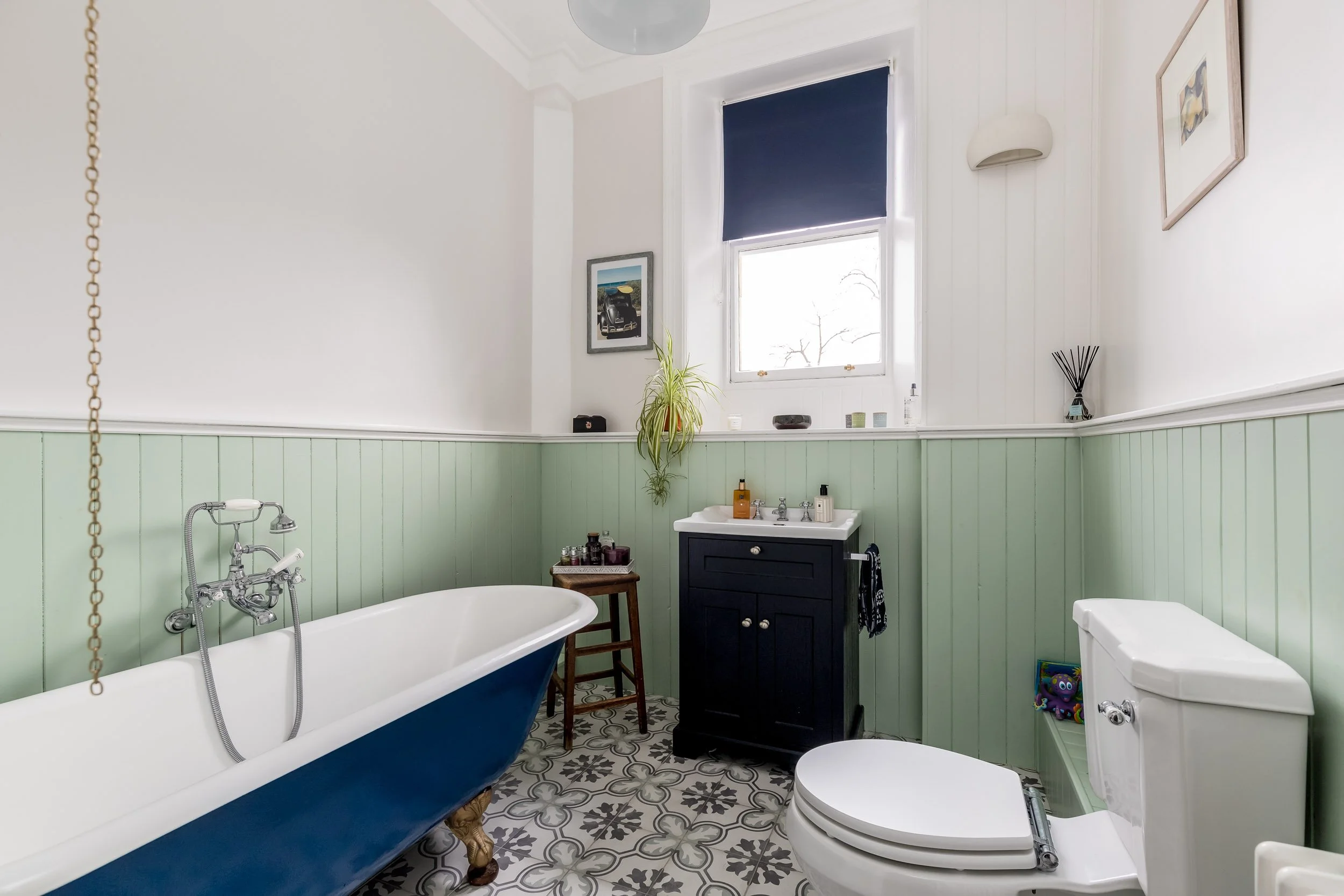 Bathroom with a clawfoot bathtub, a dark vanity with a sink, a white toilet, and patterned floor tiles, light green wainscoting, a window with a blue blind, and decorative items.