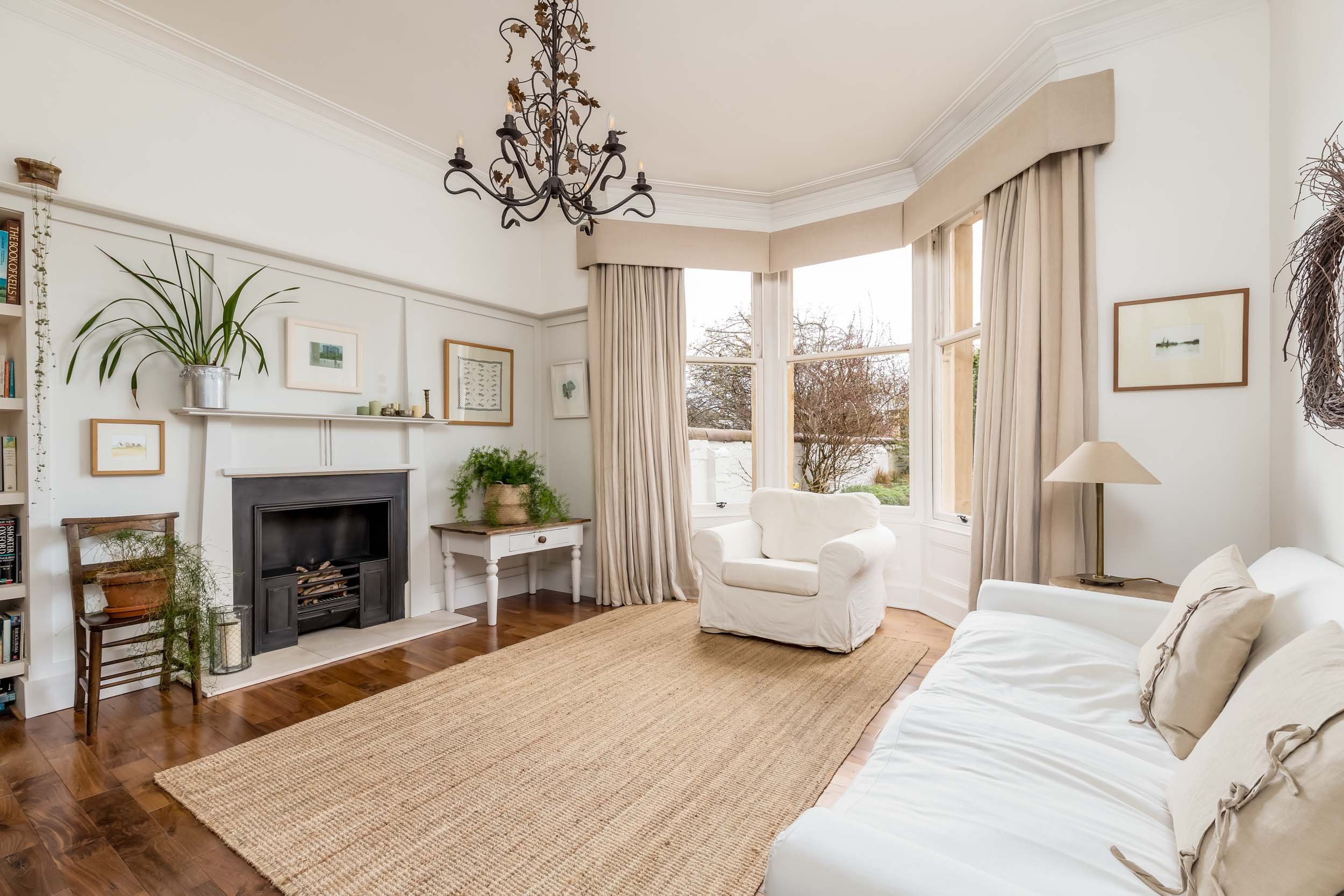 Bright living room with white walls, large bay window with beige curtains, white sofa, single armchair, fireplace, and hardwood floor with a natural fiber rug, decorated with picture frames, plants, and a black chandelier.