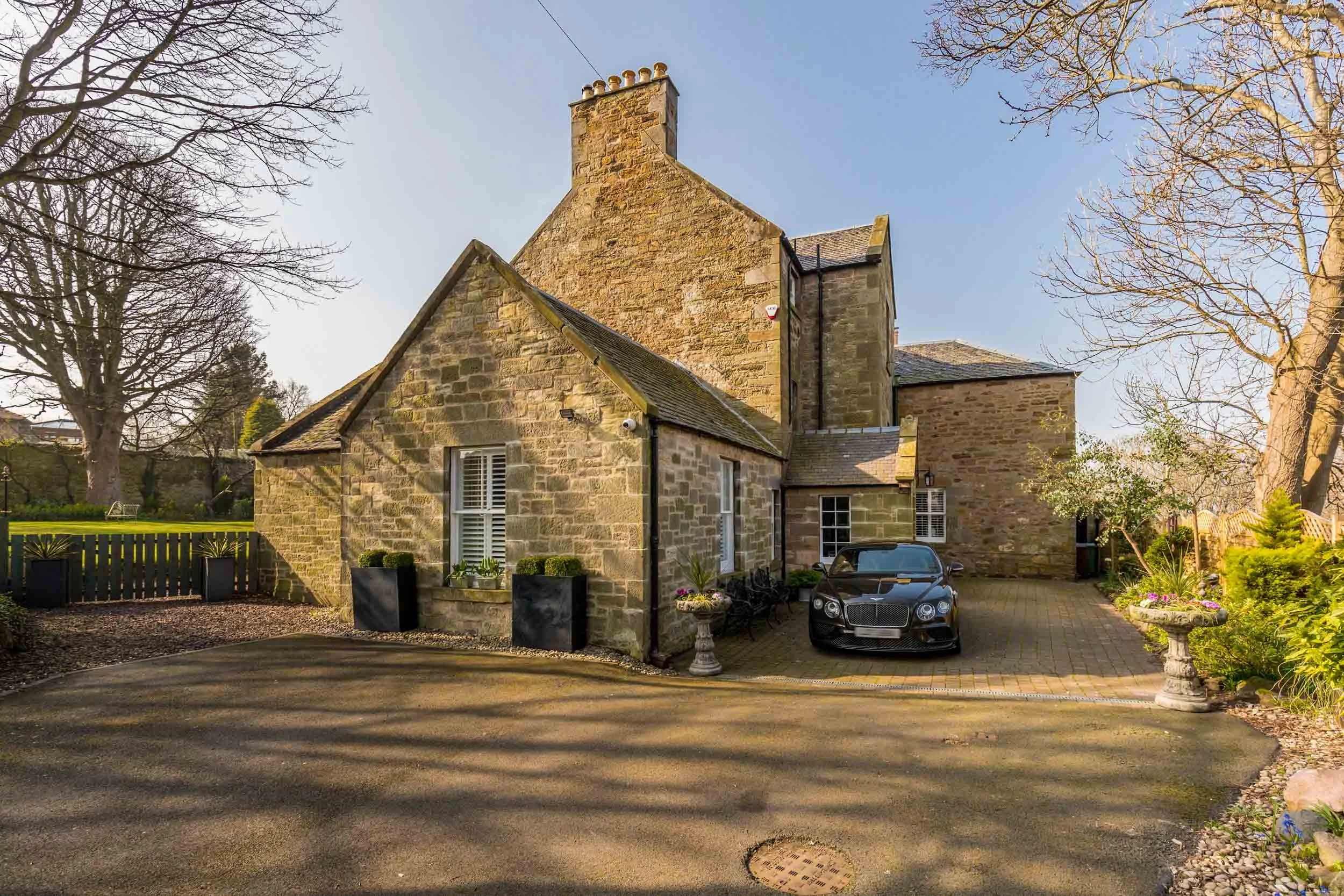 A stone house with a black sports car parked in front, surrounded by garden and trees, under a clear blue sky.