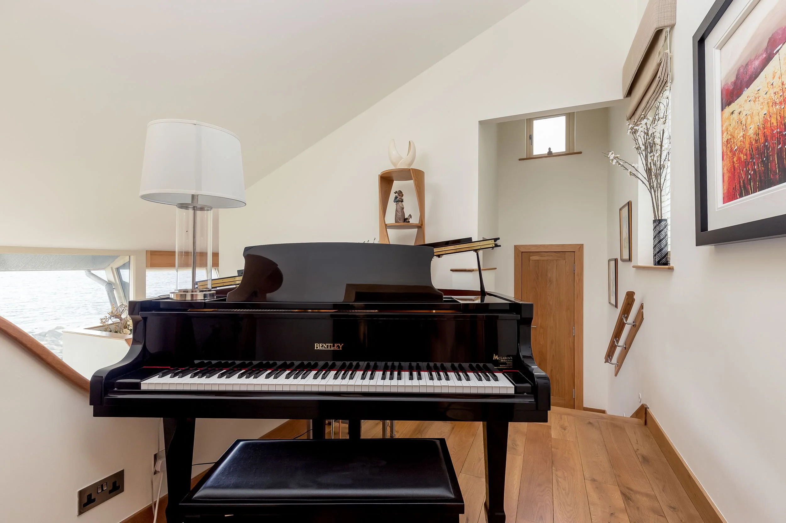 Black Bentley grand piano in a bright room with wooden floors, a table lamp, framed artwork, shelves, and a small window.
