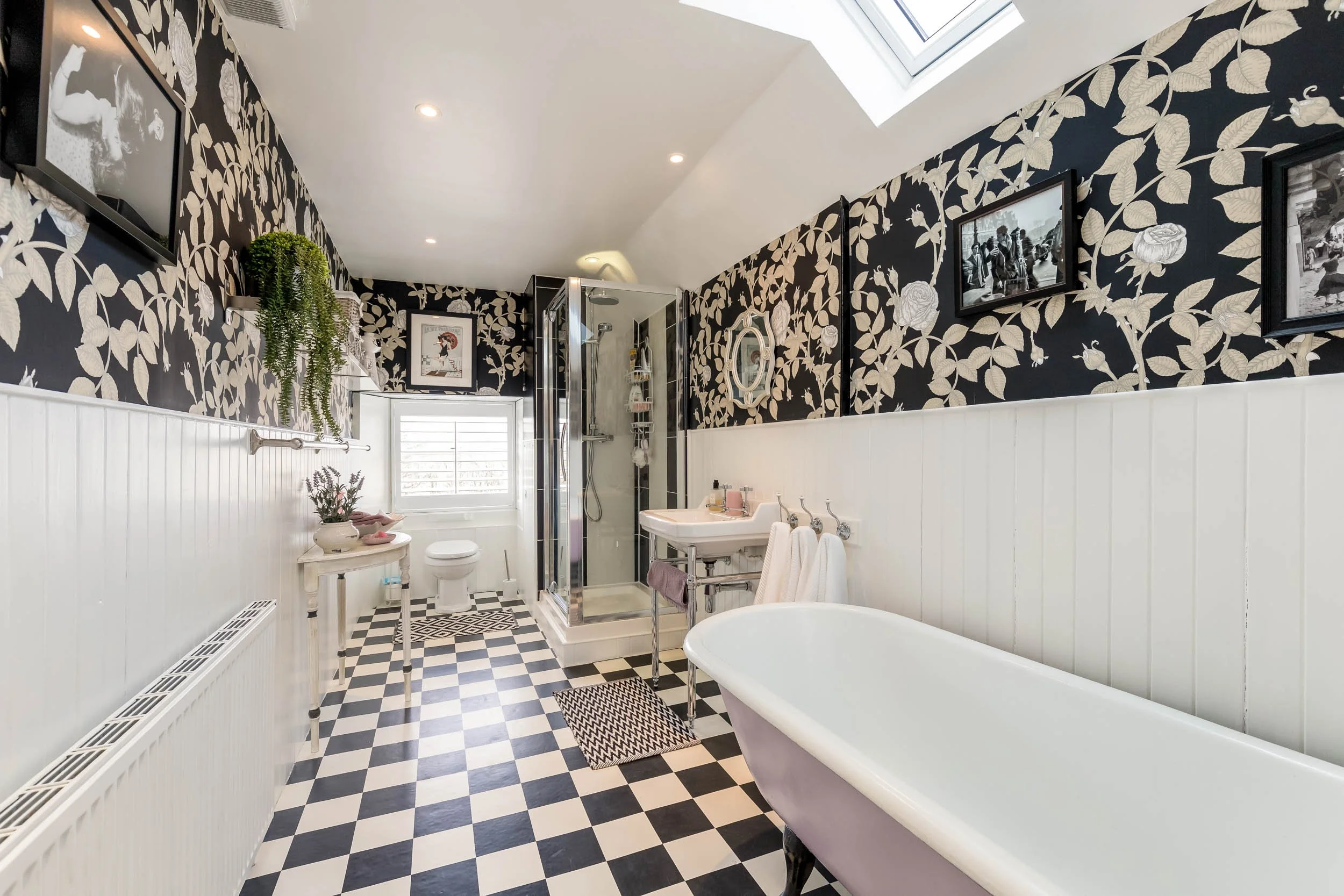 Bathroom with black and white floral wallpaper, white wainscoting, a clawfoot bathtub, shower enclosure, sink with towels, window, and framed black and white photos.