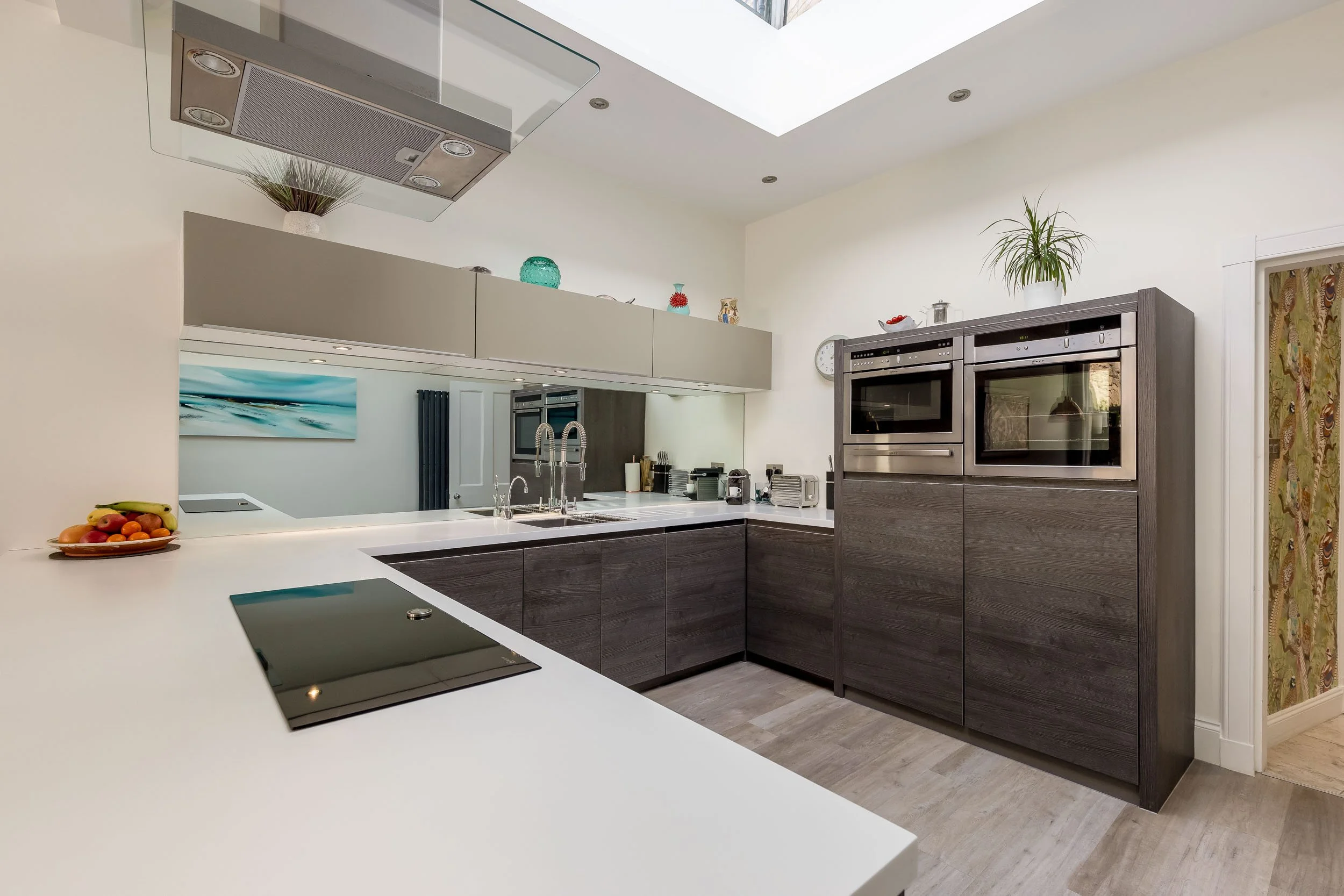 Modern kitchen with dark wood cabinets, white countertops, built-in ovens, and plants on top of cabinets and a doorway with patterned wallpaper