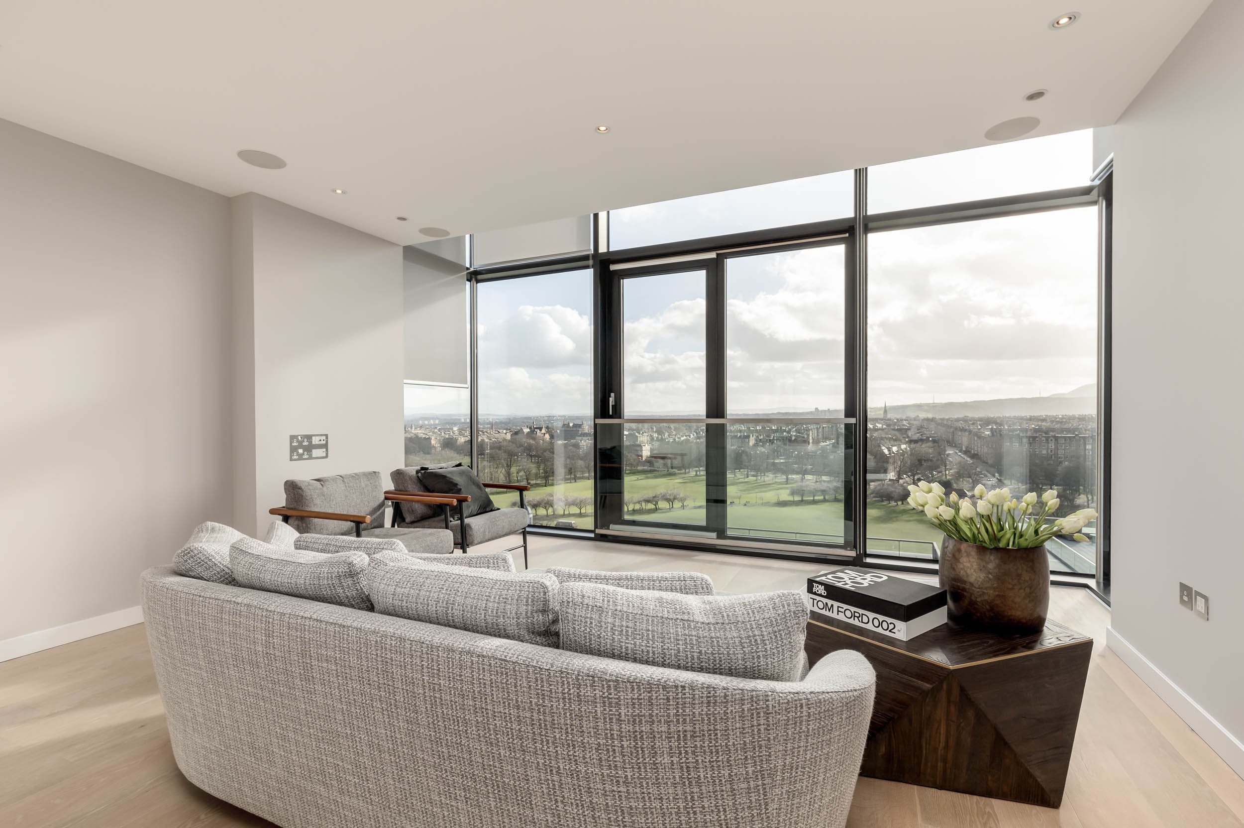 Modern living room with a large gray curved sofa, two gray armchairs, a wooden coffee table with a potted plant and books, and floor-to-ceiling windows showing a cityscape and green park outside.