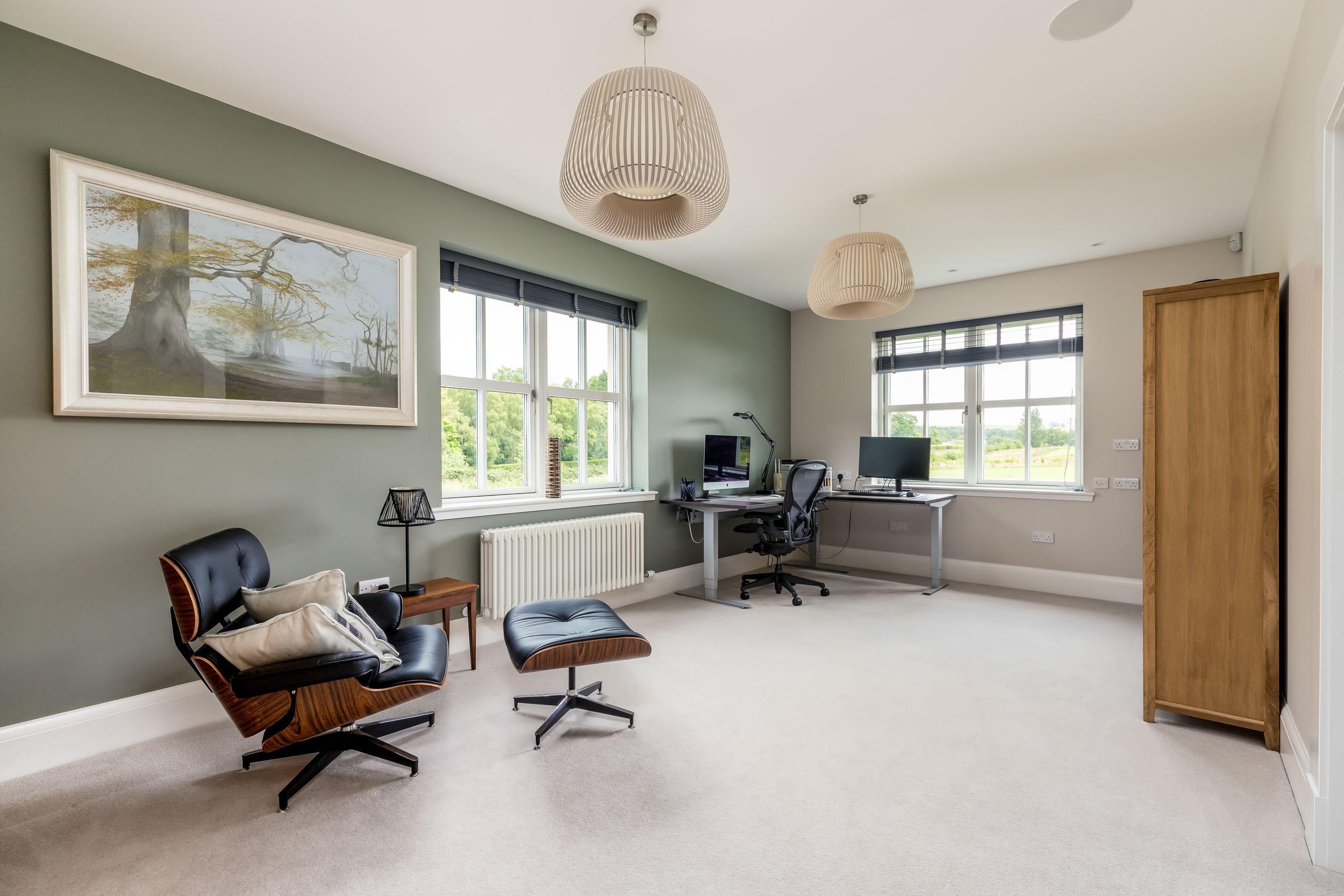 Modern home office with two desks, computer monitors, black office chairs, a lounge chair with ottoman, a side table with lamp, large windows with black blinds, framed artwork, and light fixtures, beige carpeted floor, green and white walls.