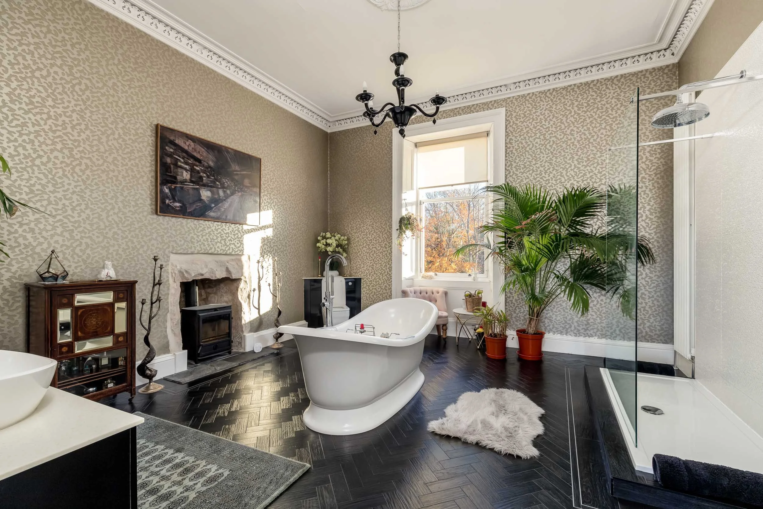 A vintage-style bathroom with a freestanding bathtub, black herringbone hardwood floors, and large window with autumn trees outside. Decor includes a chandelier, potted plants, a fireplace, and a glass shower enclosure.