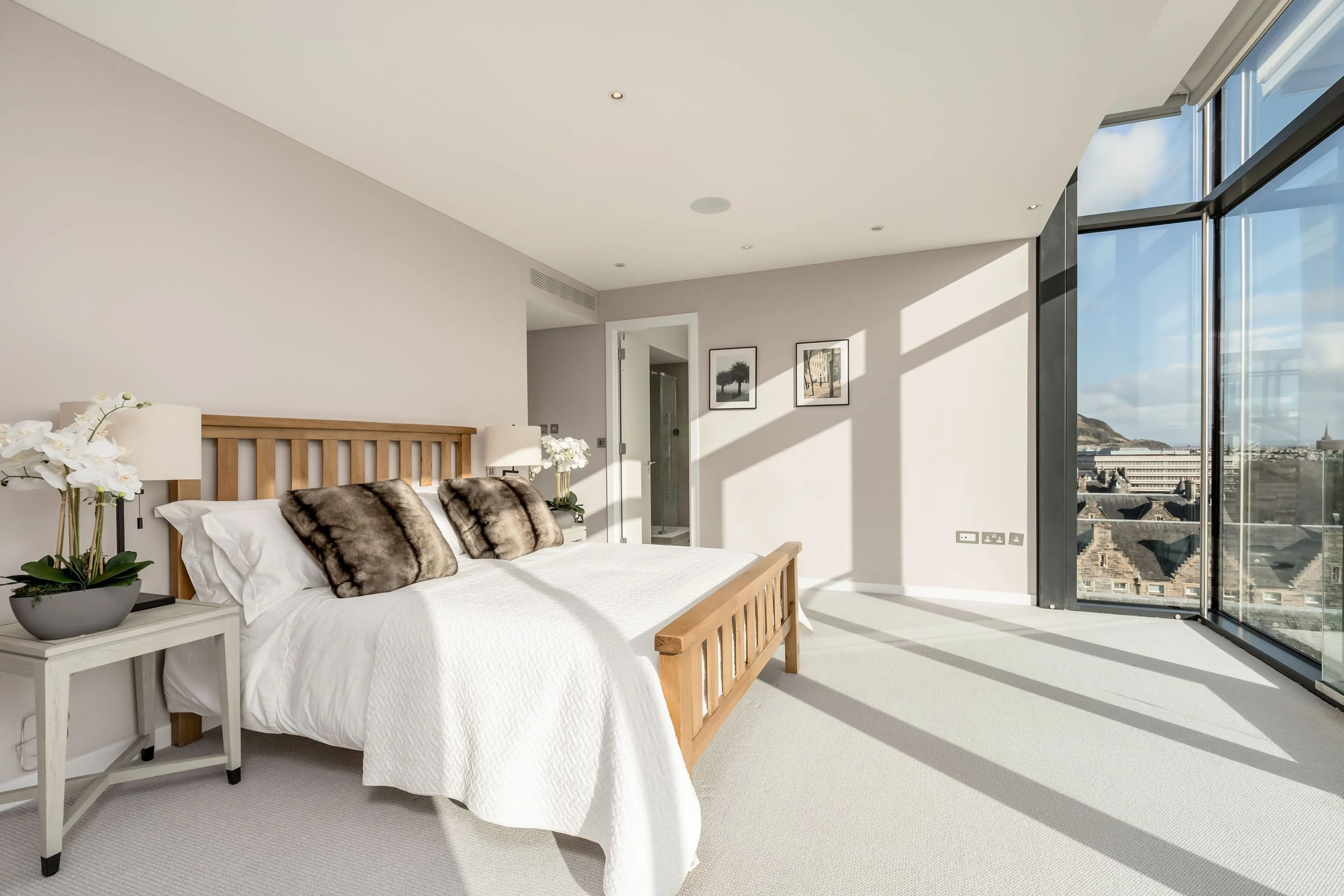 A bright, modern bedroom with a large wooden bed, white bedding, and fur pillows. Side tables with plants and lamps. Sunlight streaming through floor-to-ceiling windows showing cityscape views.
