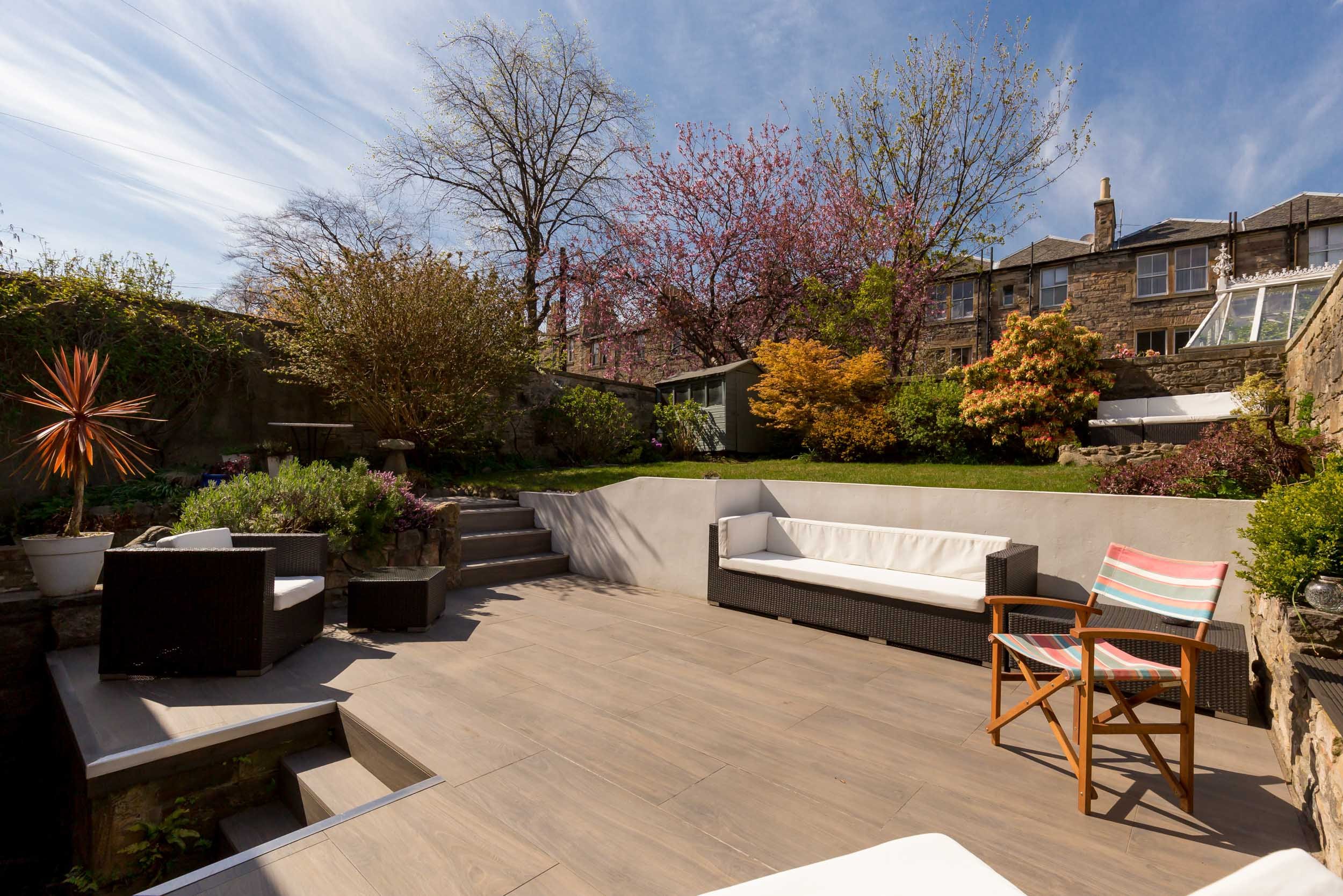 Patio area with outdoor furniture including a black wicker sofa with white cushions, a striped wooden chair, and a small black table. Surrounding the patio are planters with various plants, steps leading to a grassy yard, and a stone wall with colorf