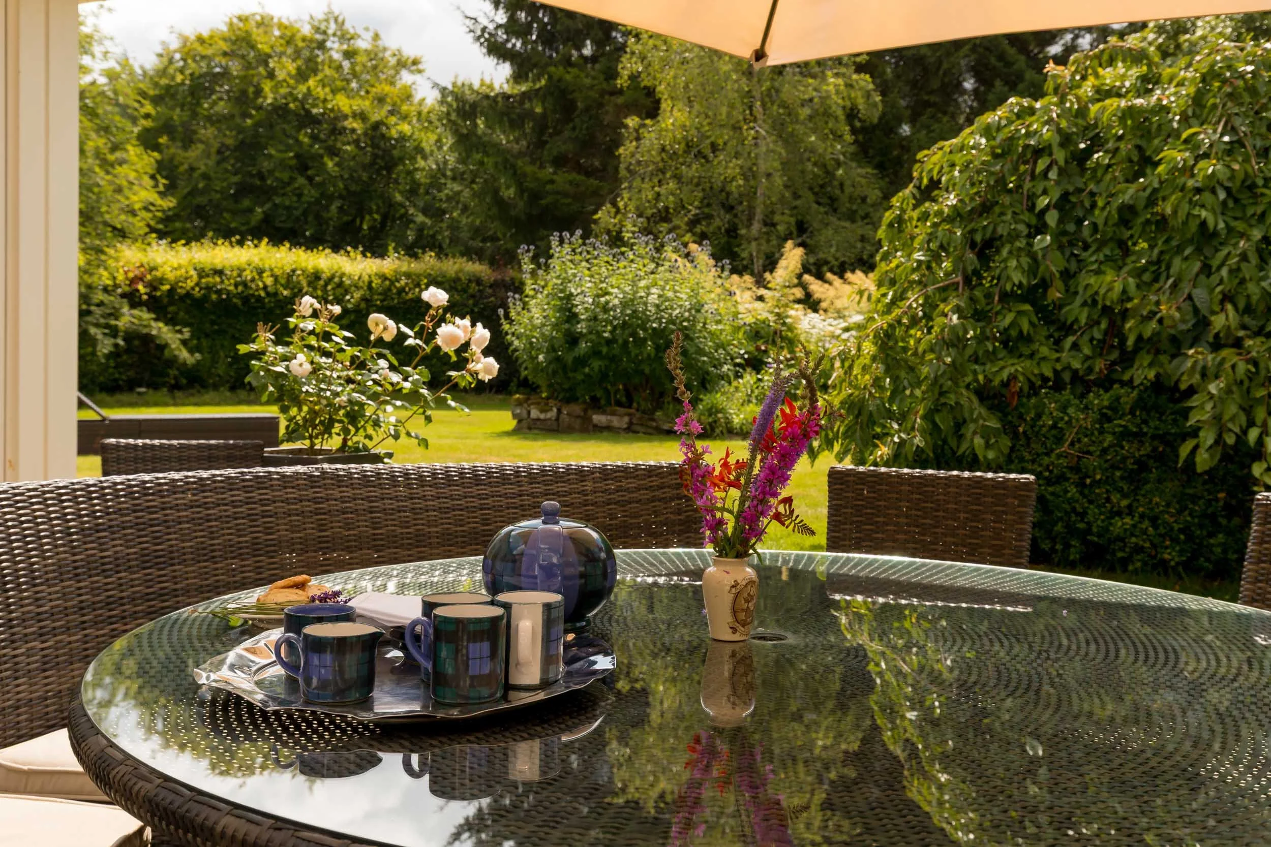 Outdoor patio table with a flower vase and cups, overlooking a lush green garden with trees and bushes on a sunny day.